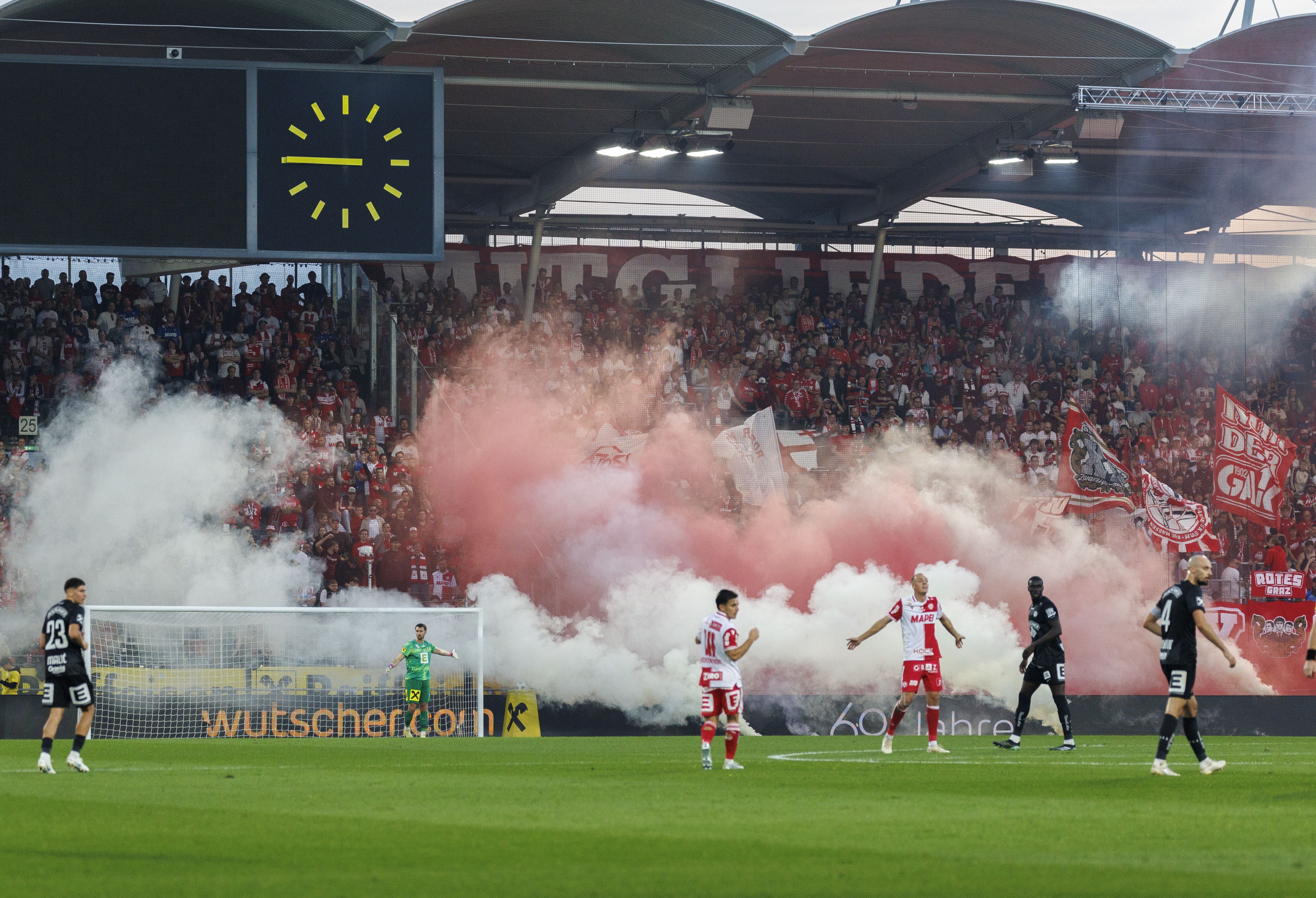 Fußballspiel mit Rauch und Pyrotechnik im Stadion, Fans jubeln auf der Tribüne.