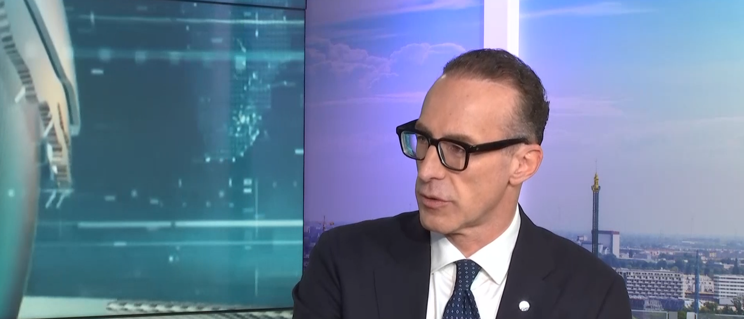 Mann im Anzug und mit Brille in einem Fernsehstudio vor Stadt- und Digitalhintergrund.