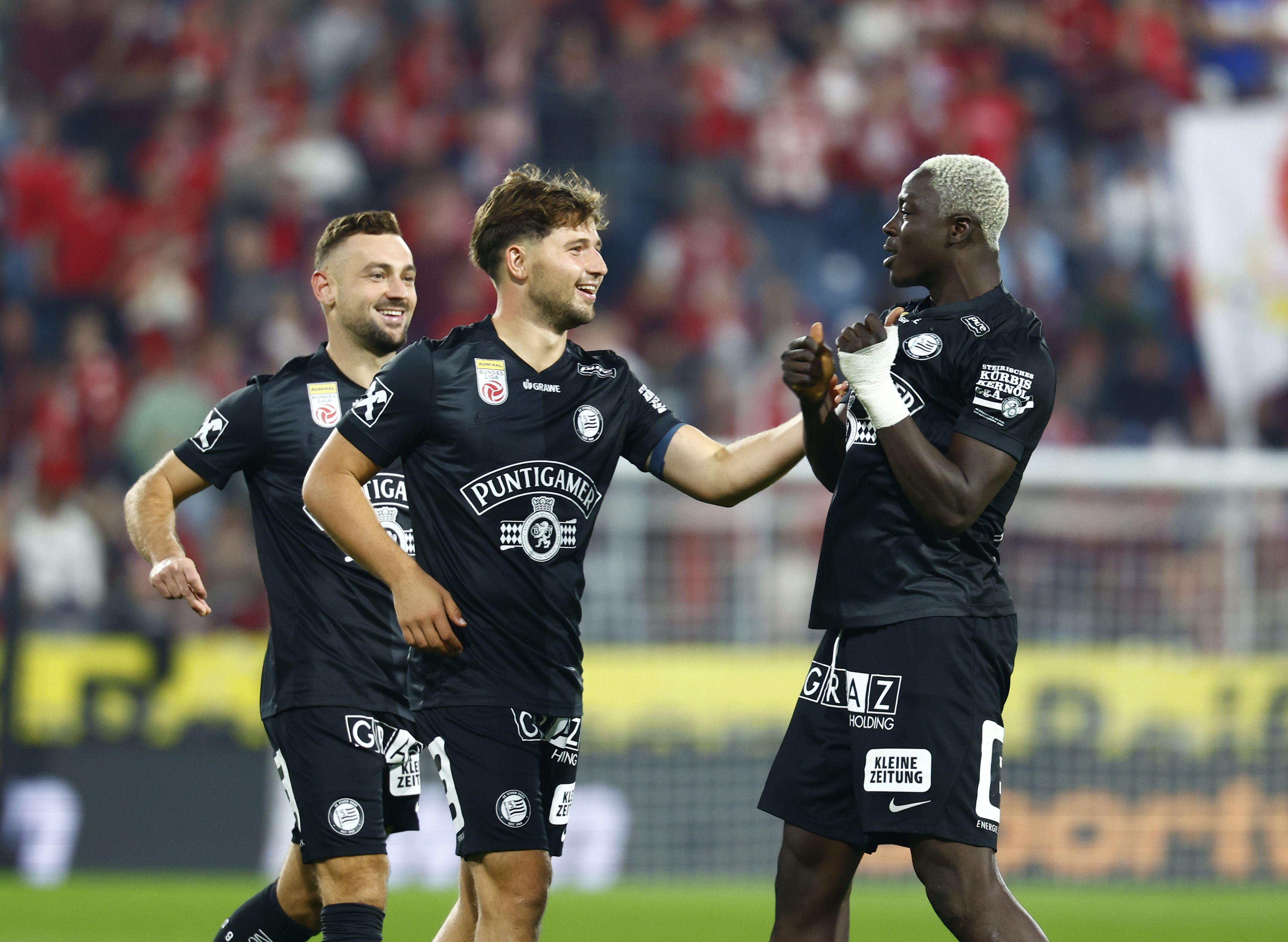 Drei Fußballer des SK Sturm Graz jubeln in schwarzen Trikots gemeinsam auf dem Spielfeld.