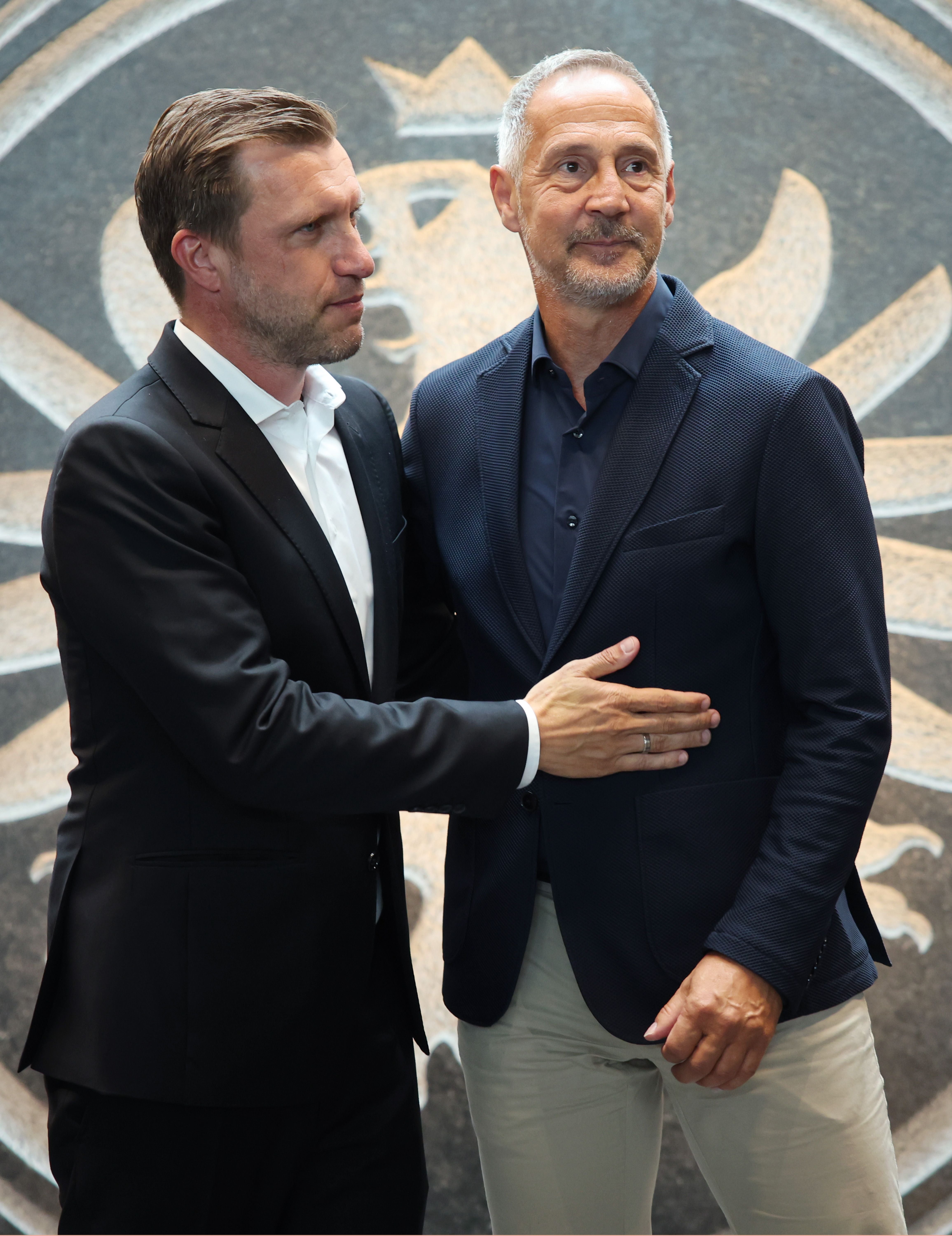 Zwei Männer in Anzügen stehen nebeneinander vor einem Wandmotiv mit Adler.