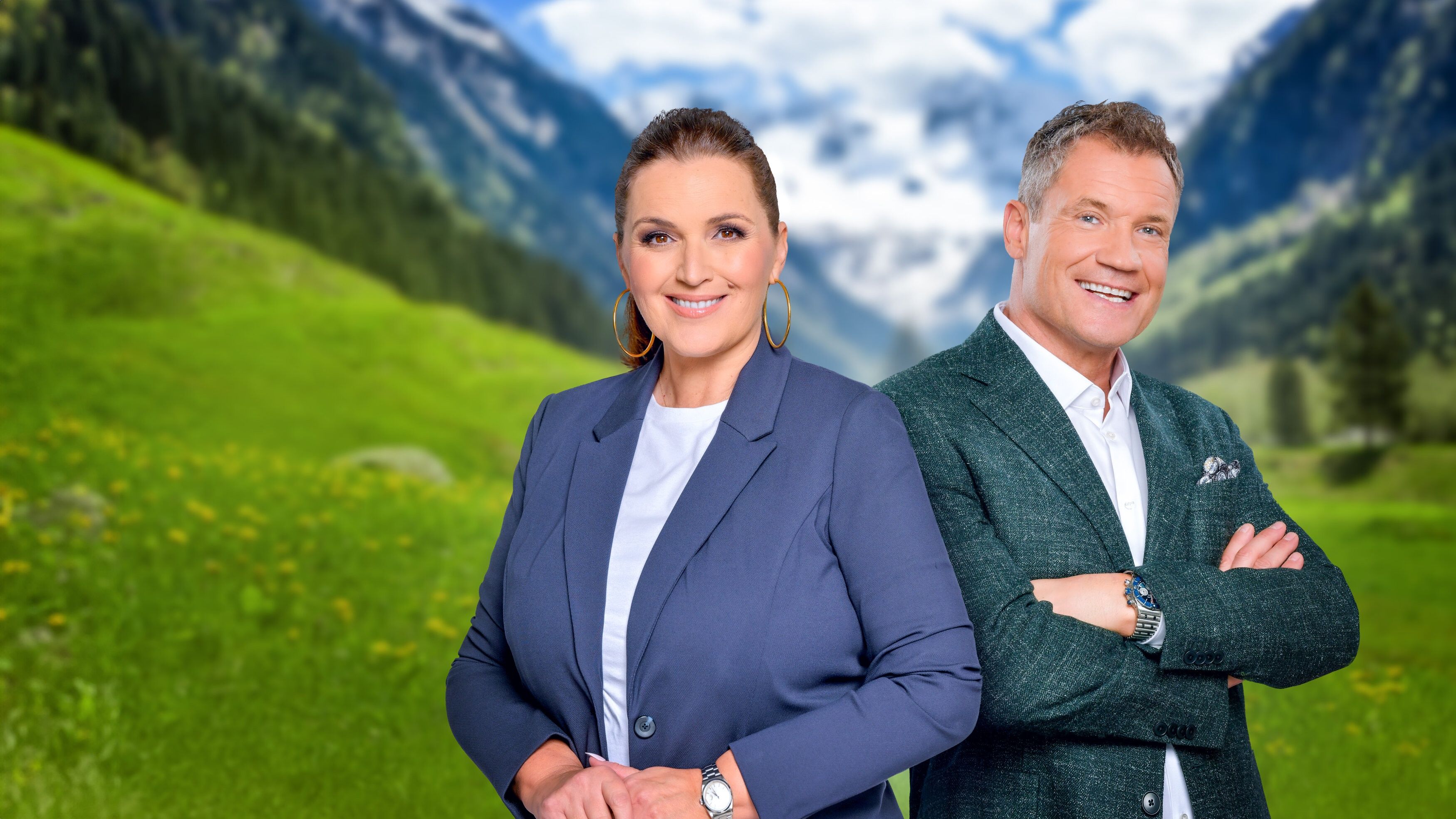 Zwei Personen stehen lächelnd vor einer grünen Berglandschaft mit verschneiten Gipfeln.