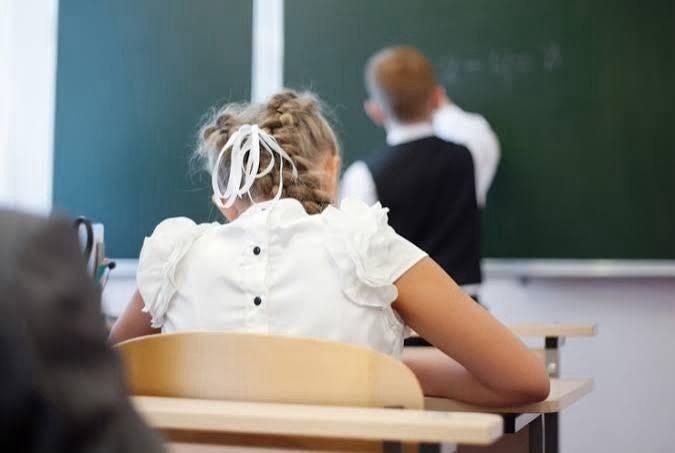 Ein Mädchen sitzt im Klassenzimmer, ein Junge schreibt an die Tafel.