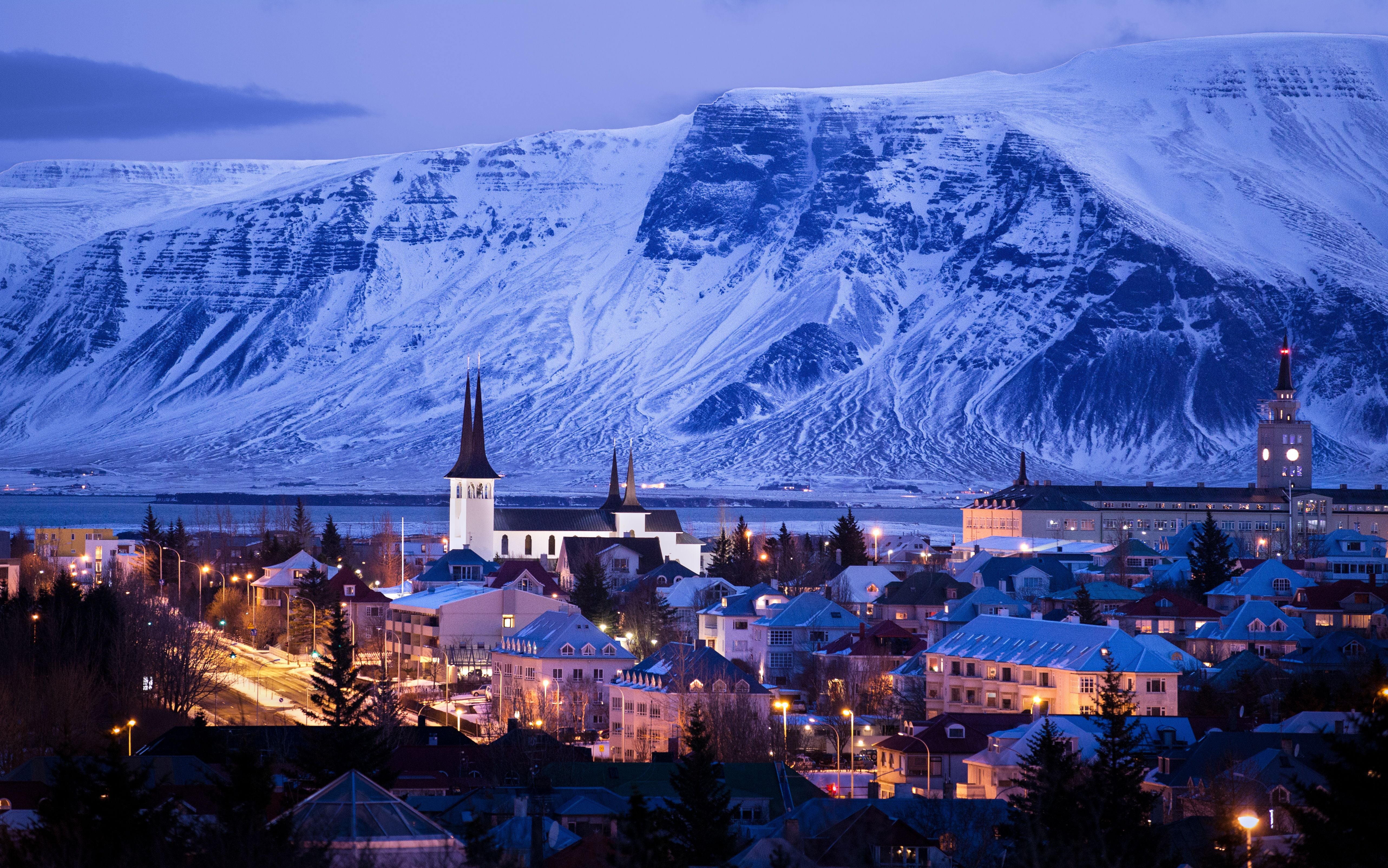 Abendliche Stadtansicht mit beleuchteten Häusern vor schneebedeckten Bergen.