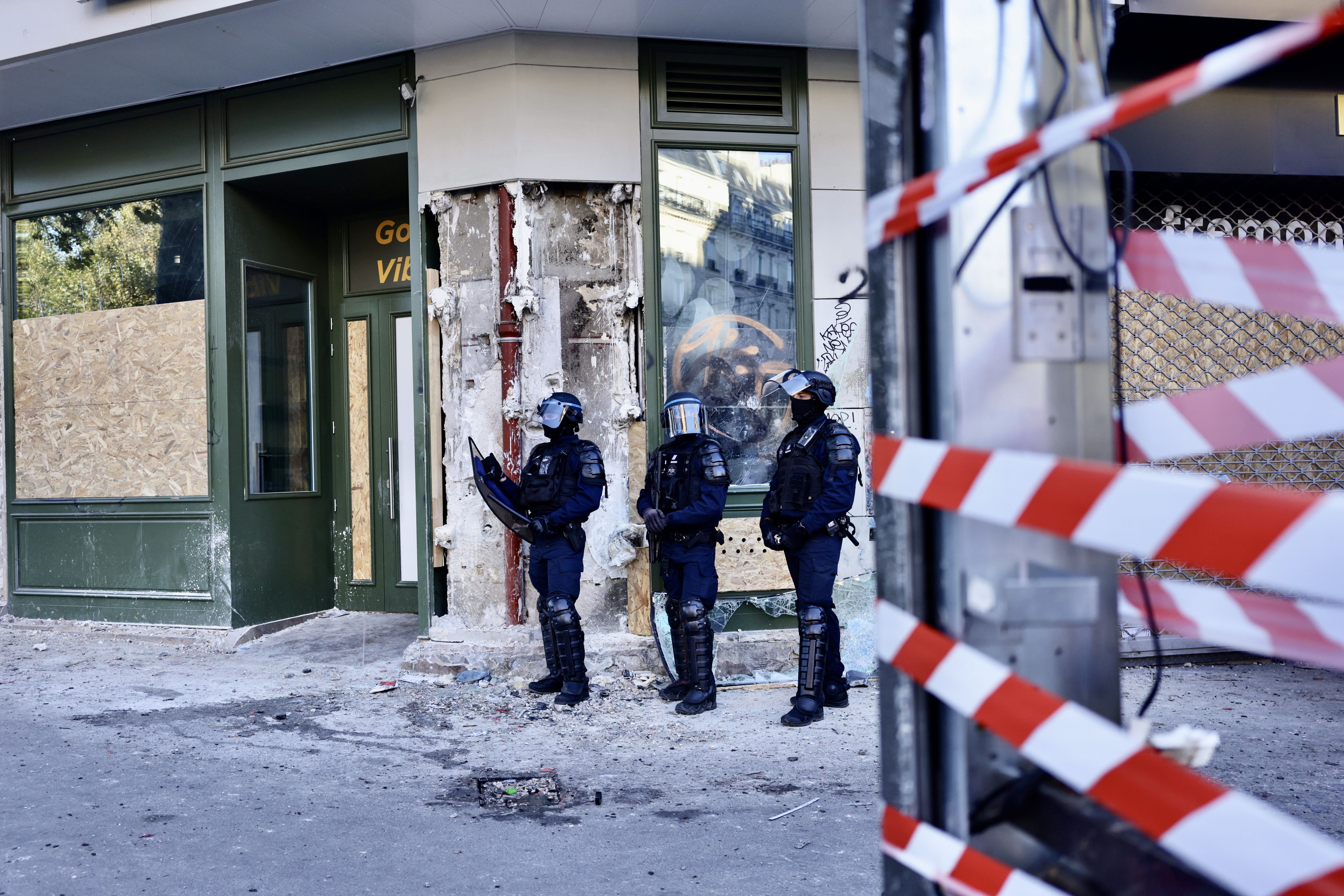 Drei Polizisten in voller Ausrüstung stehen vor einem beschädigten Gebäude, Absperrband im Vordergrund.
