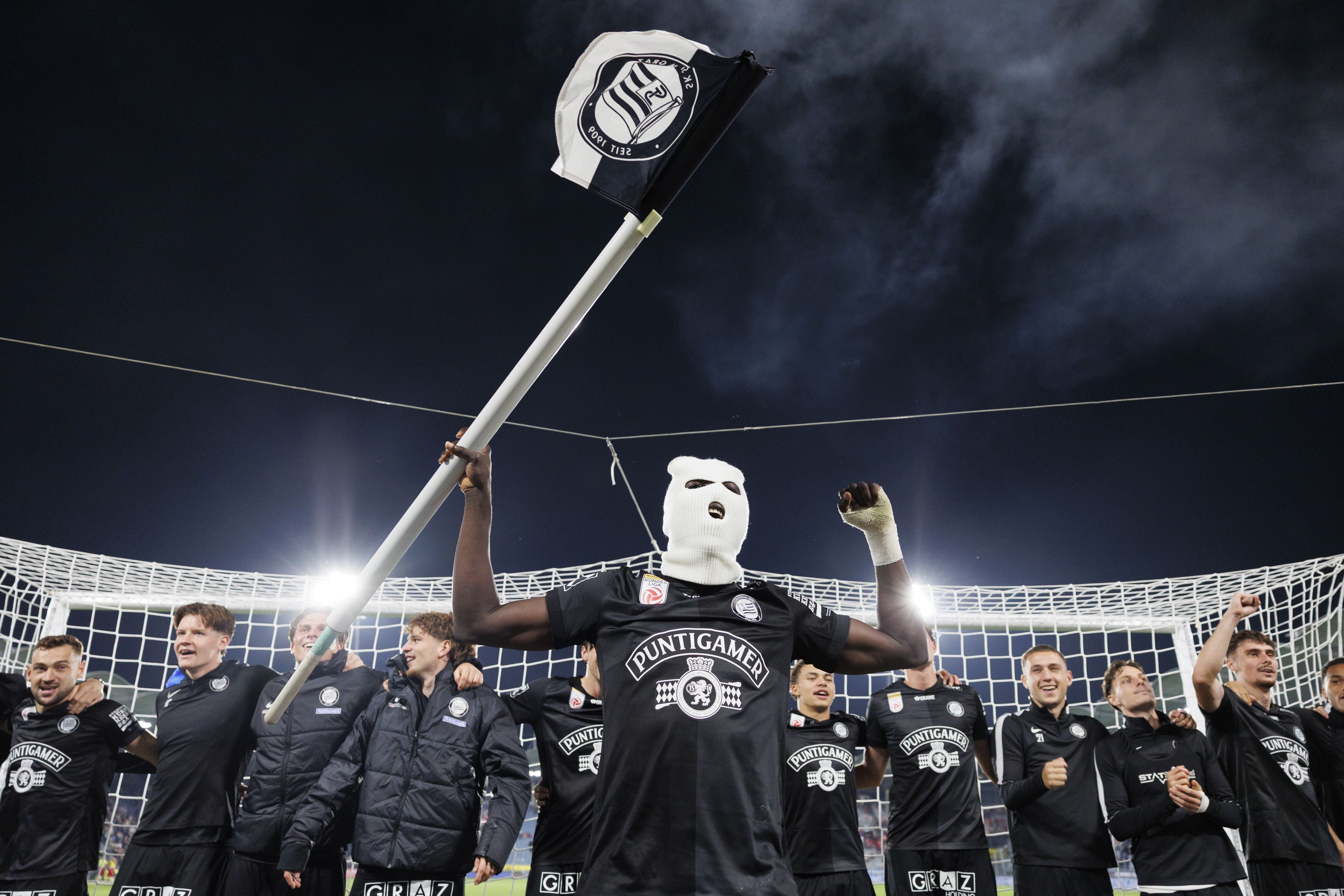 Fußballspieler von Sturm Graz feiern im Stadion, einer trägt eine weiße Sturmhaube und hält eine Fahne.