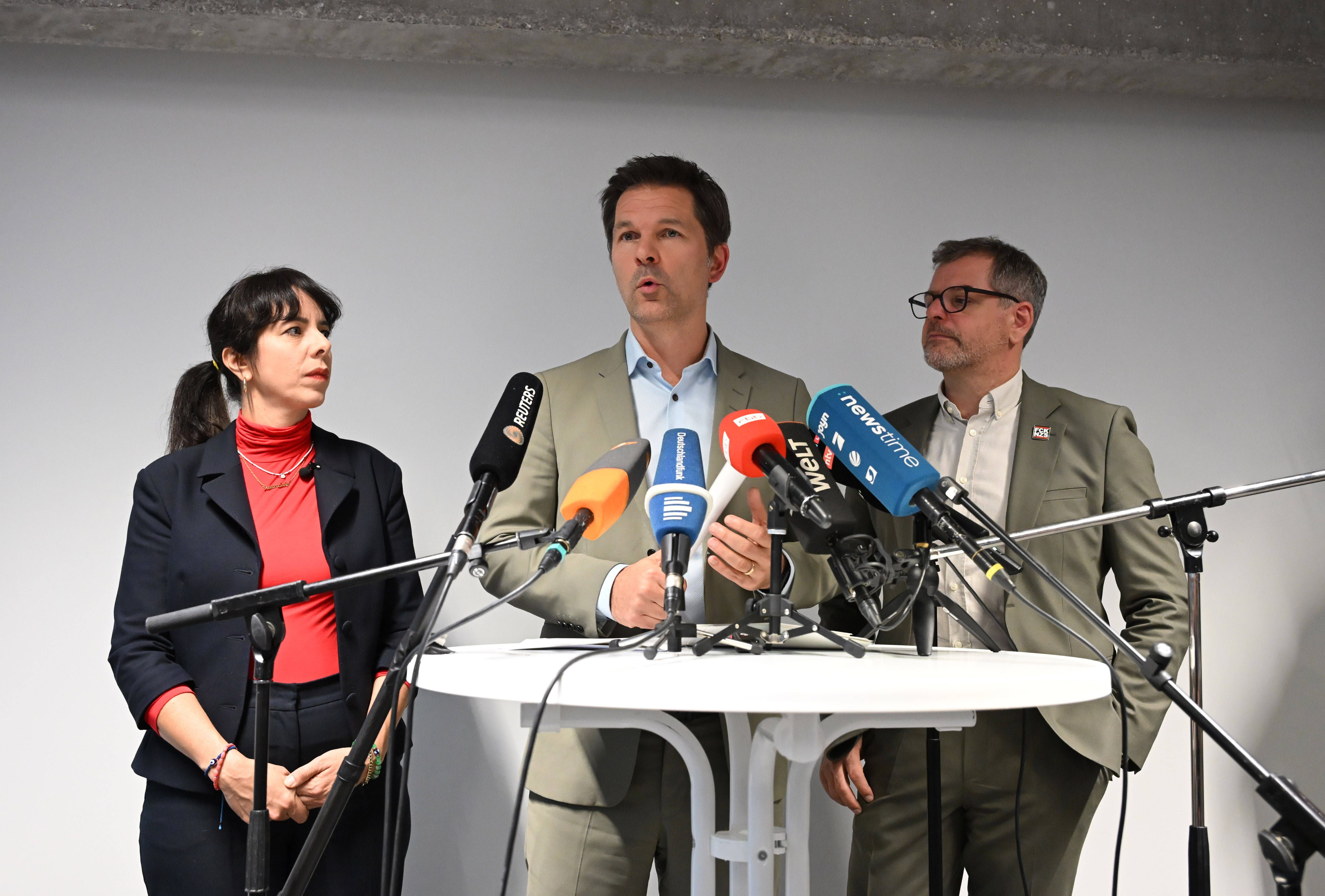 Drei Personen halten eine Pressekonferenz hinter einem weißen Tisch mit mehreren Mikrofonen.