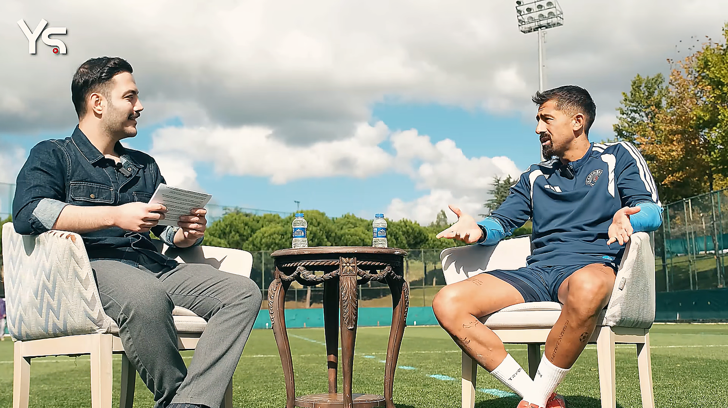 Kerem Demirbay sitzt im Trainingsdress mit ausgebreiteten Händen im Interview gegenüber von Yağız Sabuncuoğlu auf einem Sportplatz.