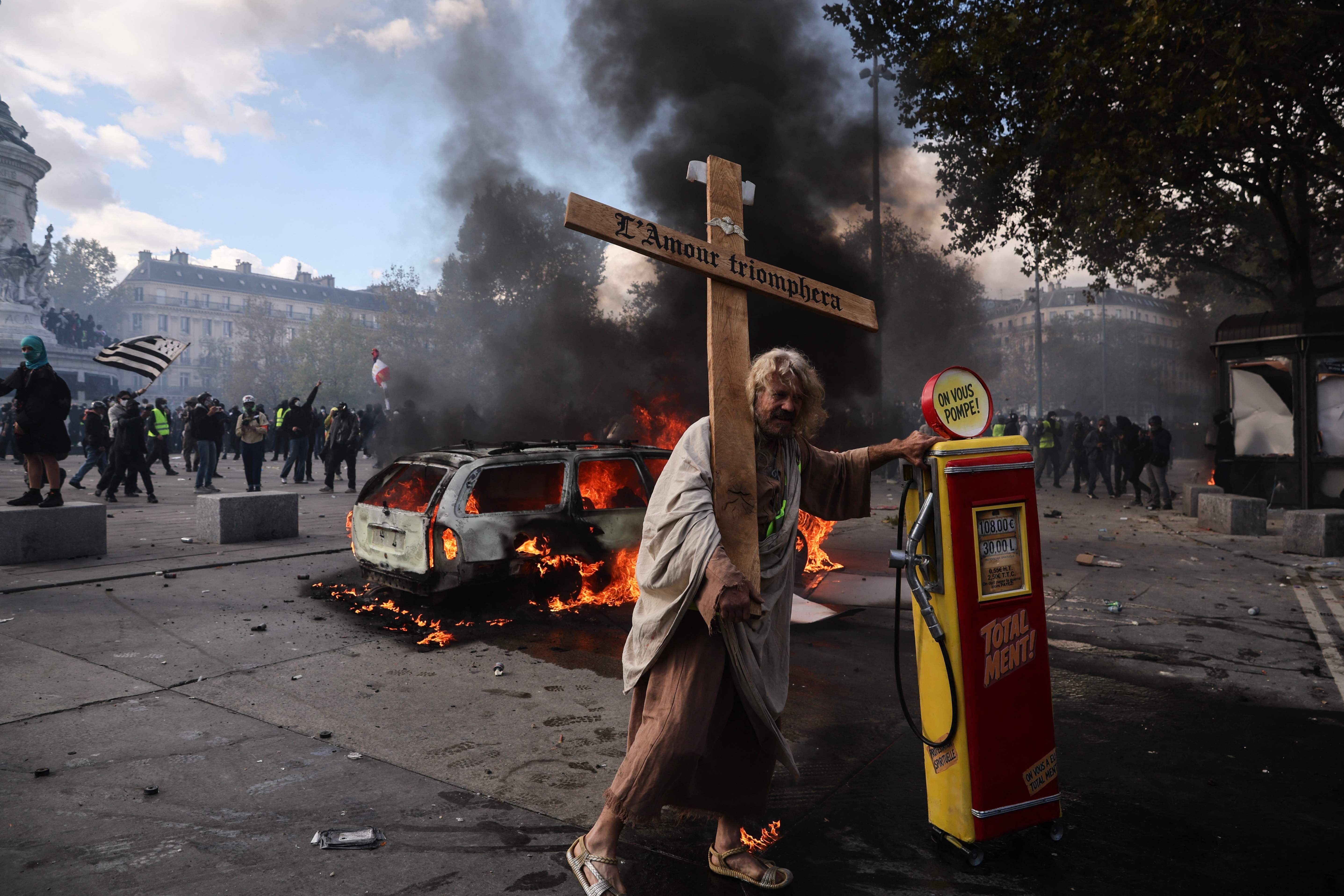 Ein Demonstrant mit Kreuz steht vor einer brennenden Auto und Zapfsäule bei Protesten in Paris.