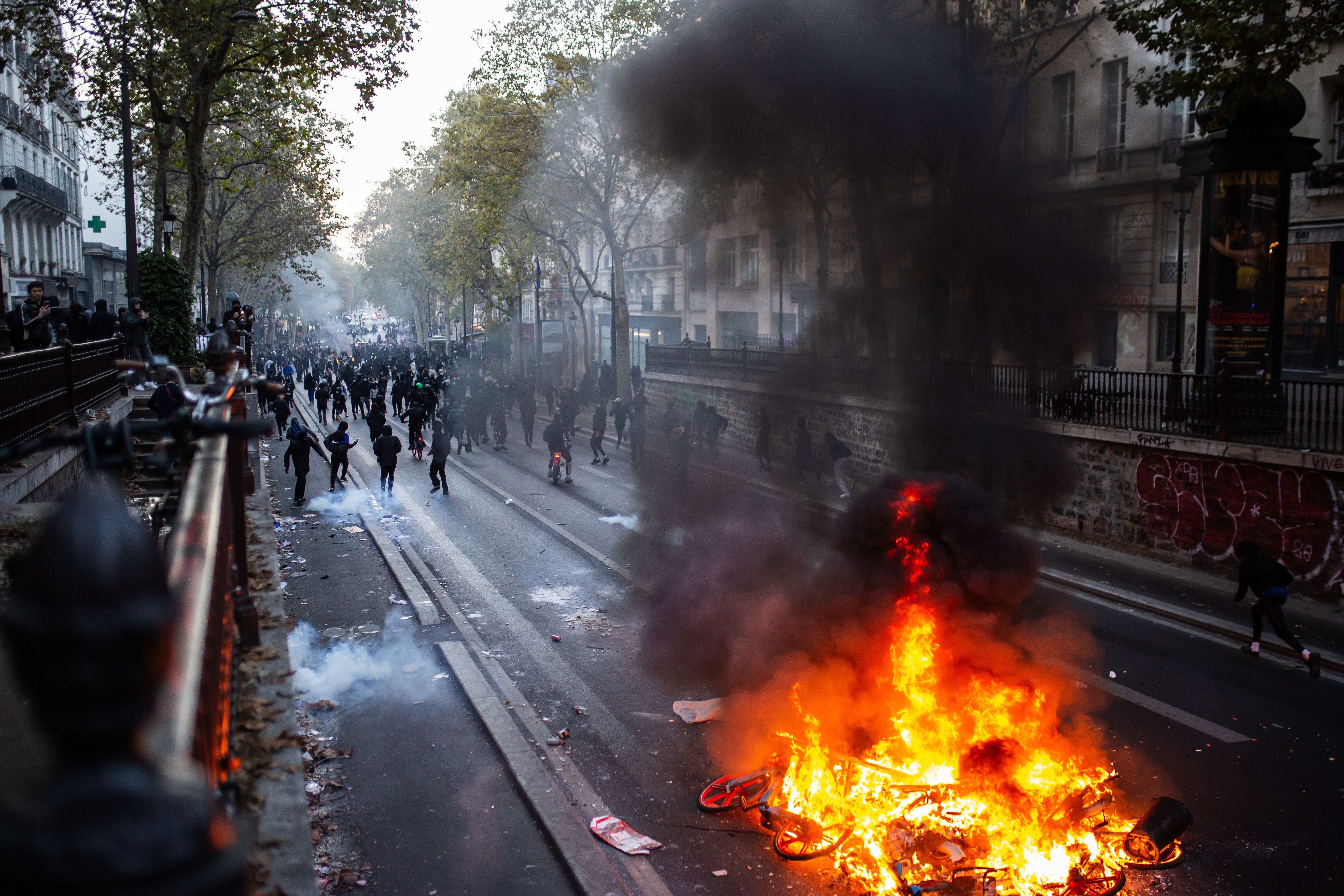 Brennende Barrikade auf einer Straße in Paris, Menschen und Rauch im Hintergrund bei Protesten.
