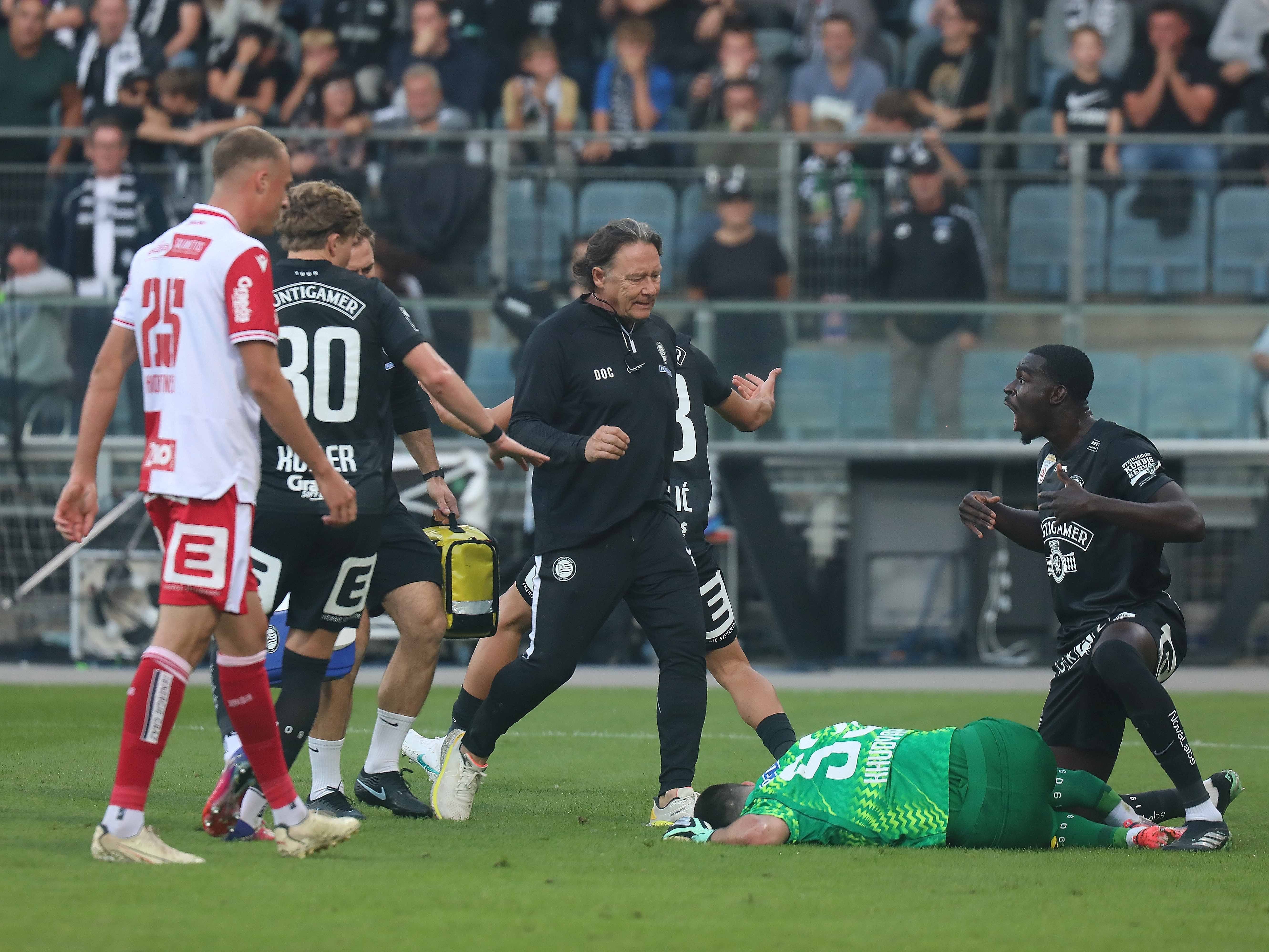 Mehrere Fußballspieler und ein Trainer reagieren auf einen am Boden liegenden Torwart während eines Spiels.
