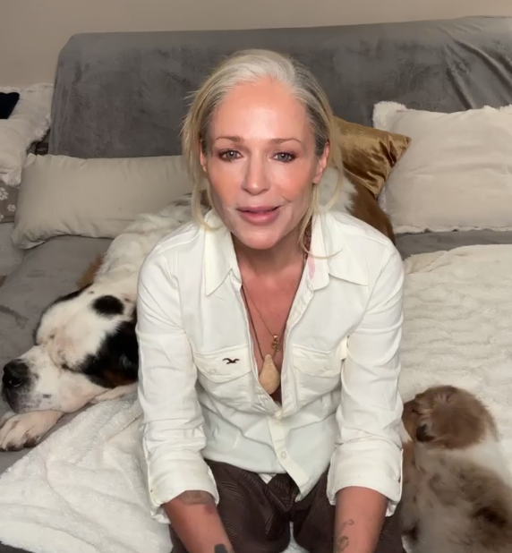Frau sitzt auf einem Bett mit einem schlafenden Hund und einem kleinen Welpen neben sich.