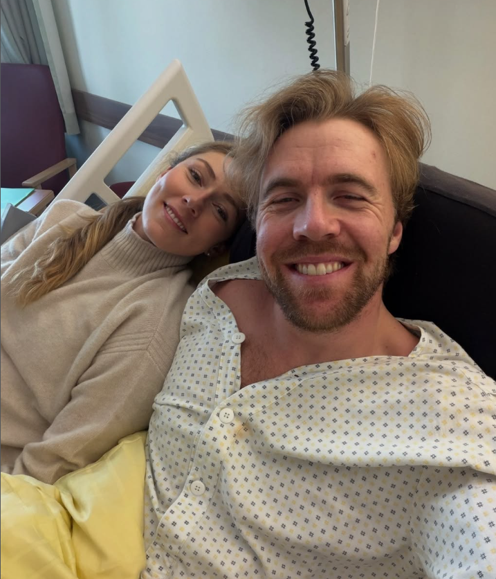 Ein Mann im Krankenhausbett und eine Frau sitzen lächelnd eng zusammen für ein Selfie.