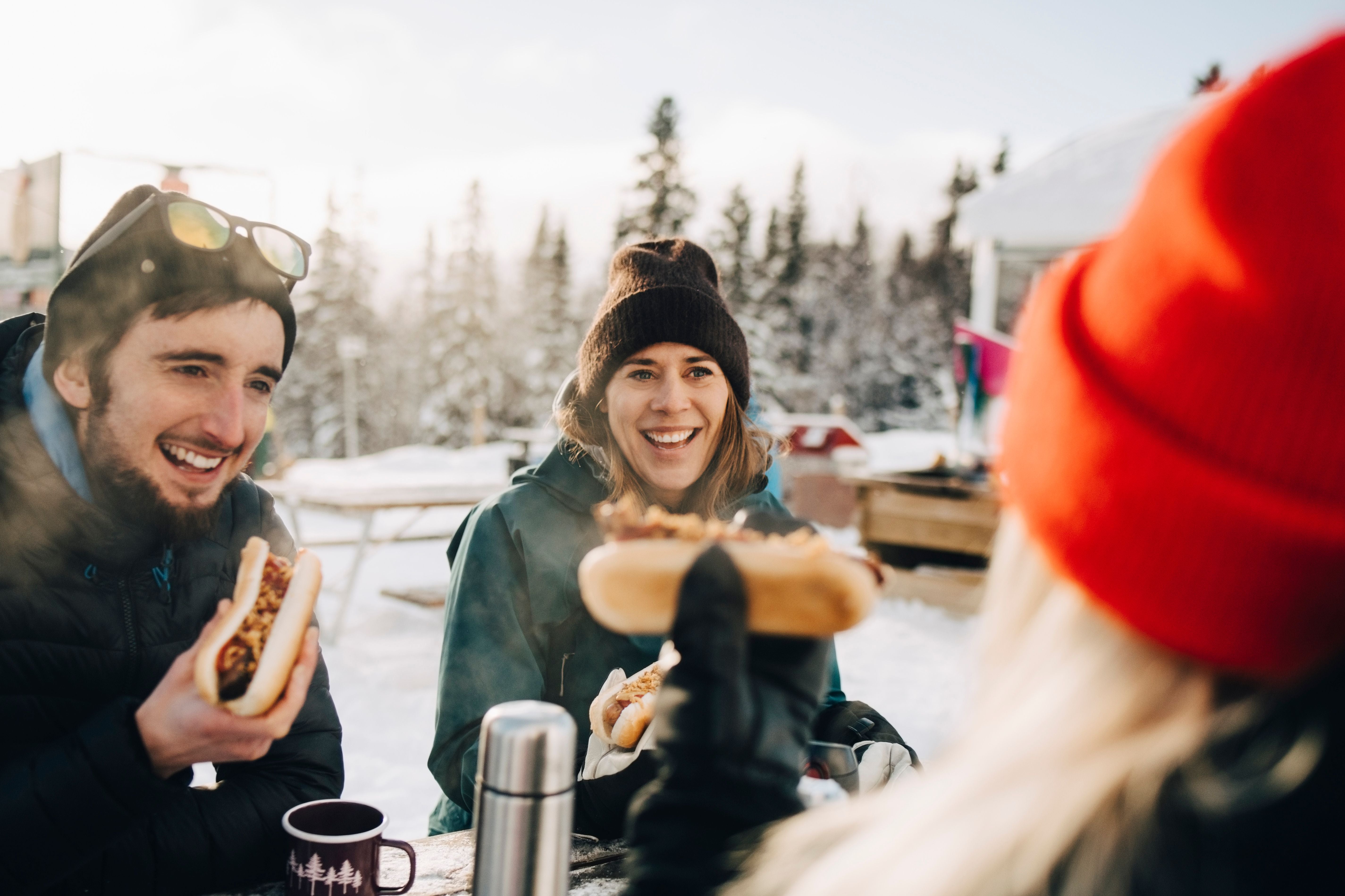 Drei lachende Menschen essen Hotdogs im Winter draußen im Schnee, mit heißen Getränken.