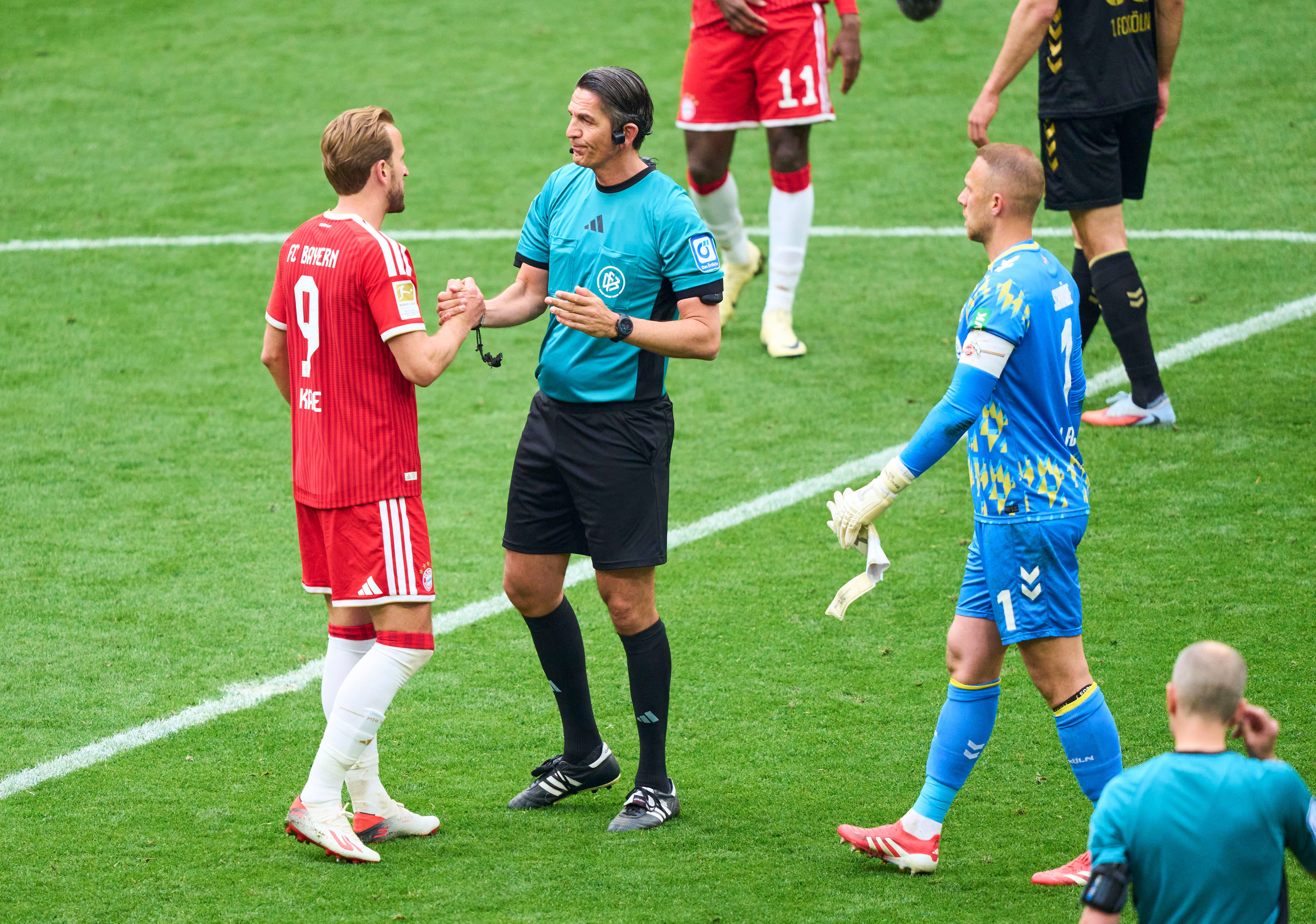 Schiedsrichter begrüßt einen Bayern-Spieler und einen Torwart nach Abpfiff auf dem Fußballfeld.