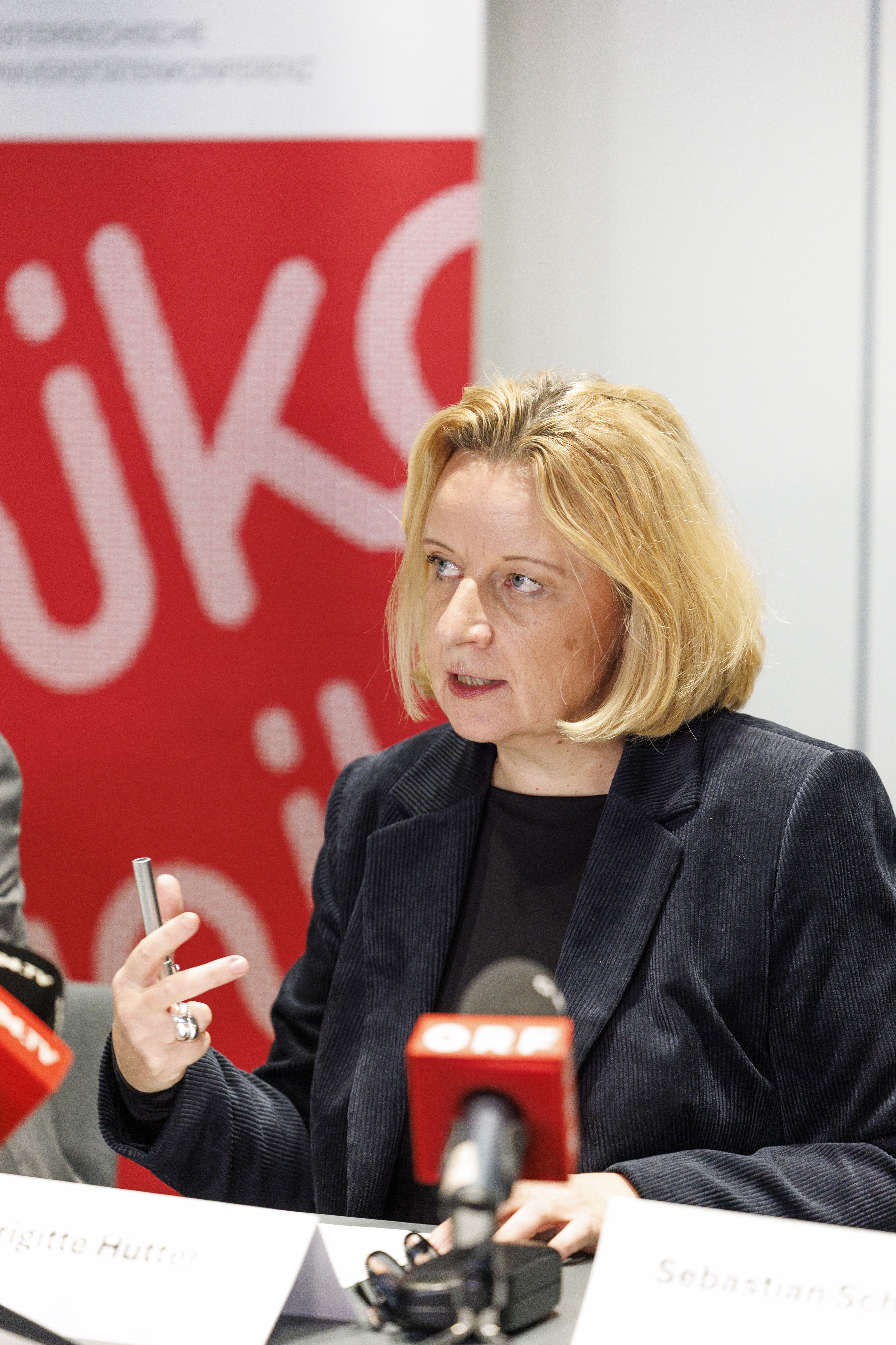 Eine Frau spricht bei einer Pressekonferenz, Mikrofone und Namensschild vor ihr.