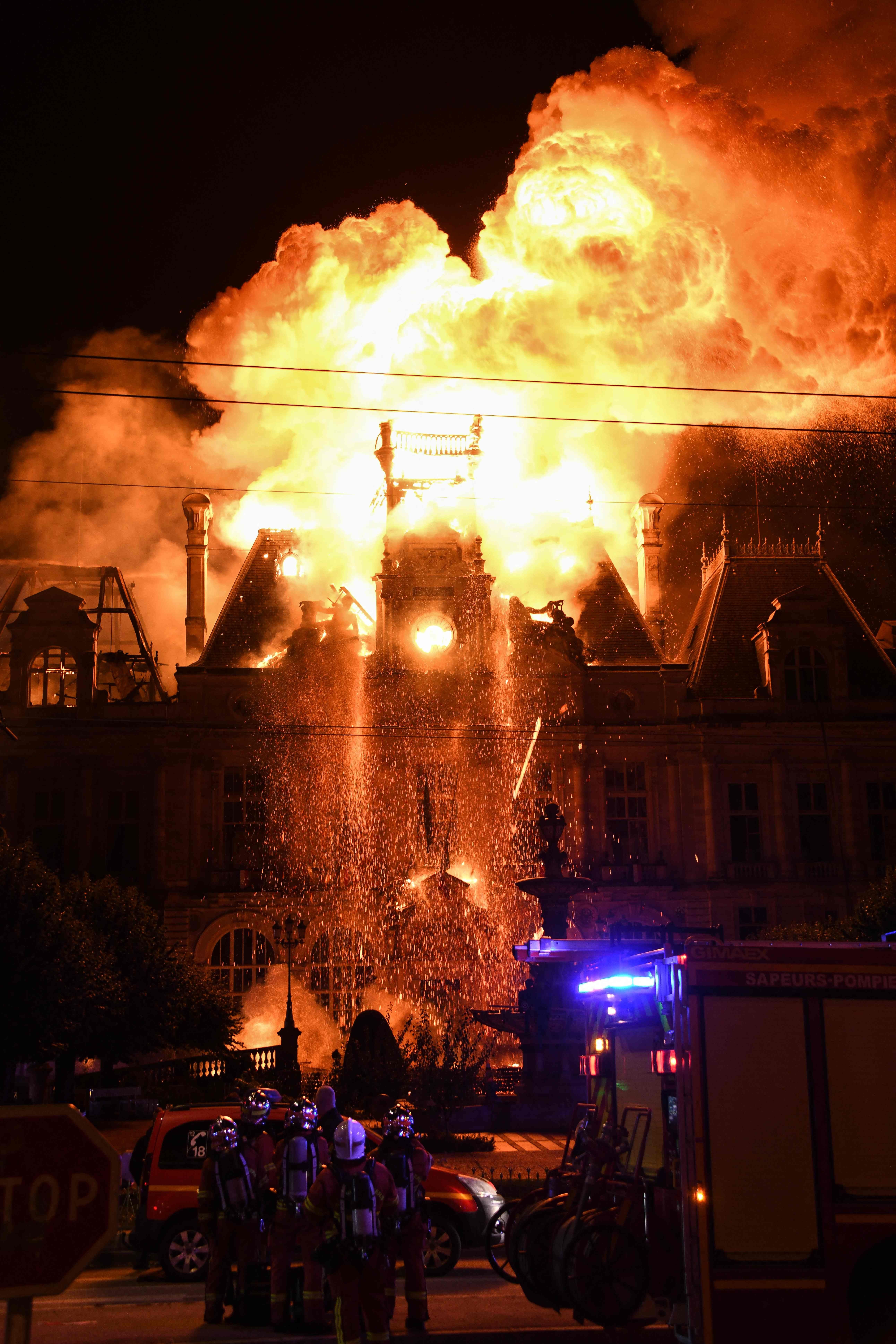 Großes Feuer zerstört ein historisches Gebäude, Feuerwehrleute stehen davor, Flammen lodern am Dach.