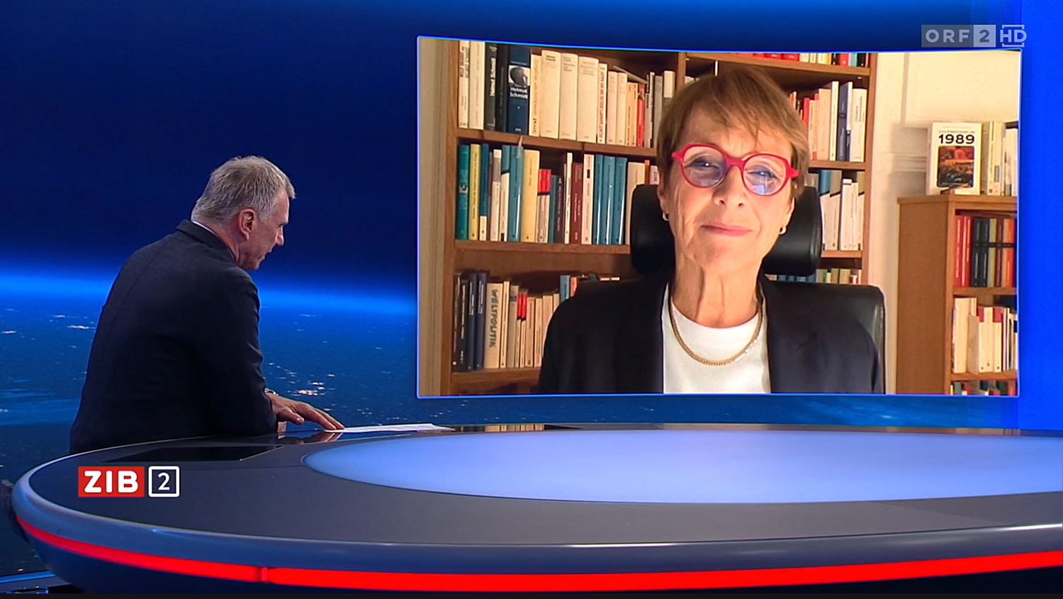 Fernsehinterview mit einer Frau auf einem großen Bildschirm und einem Moderator im Studio.