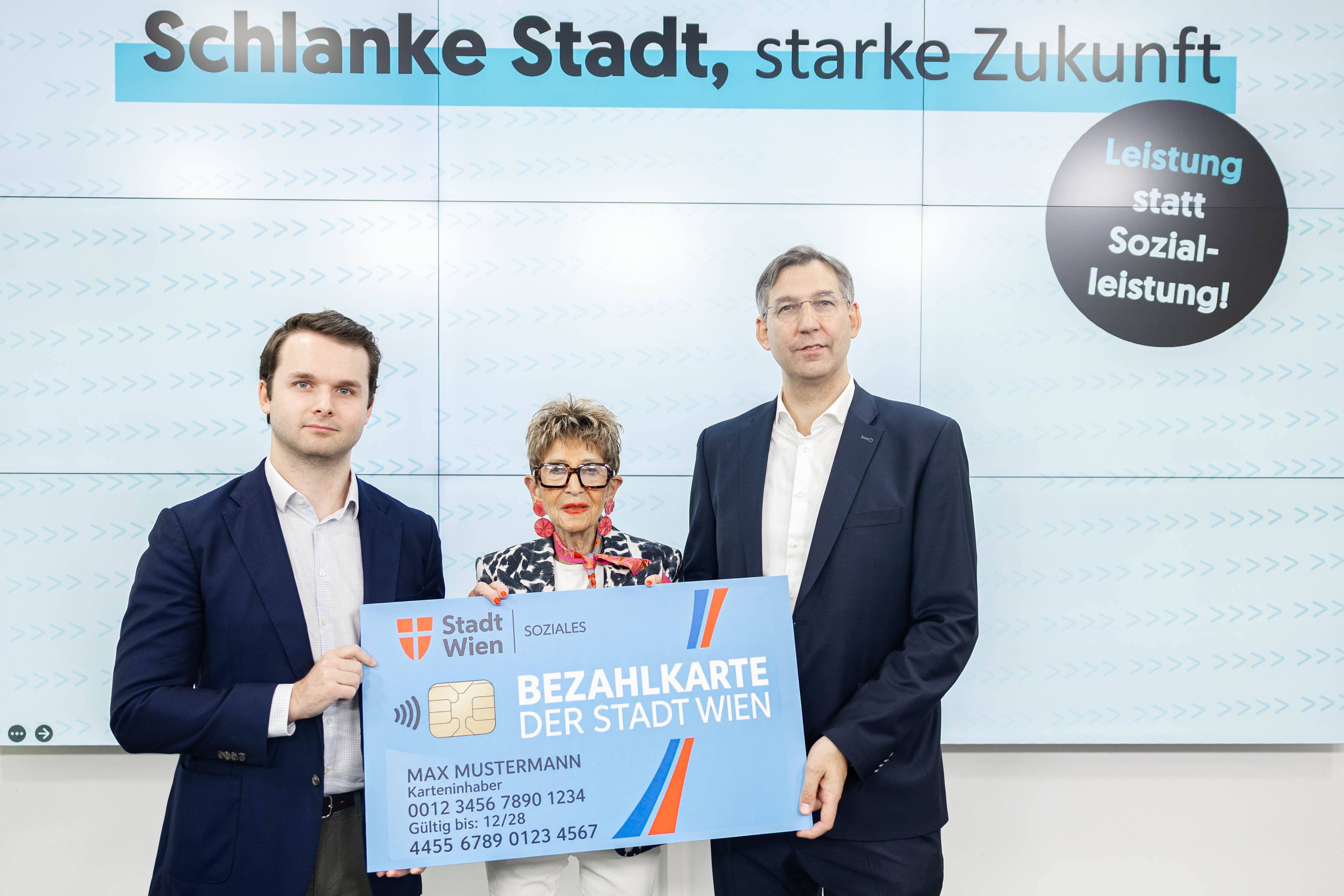 Drei Personen halten eine große Bezahlkarte der Stadt Wien vor einem Präsentationsbildschirm.