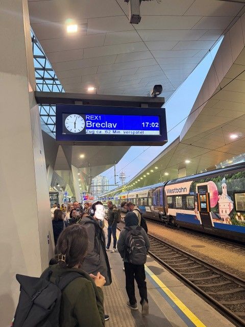Menschen warten am Bahnsteig, darüber eine Anzeigetafel für den Zug nach Breclav mit Verspätung.