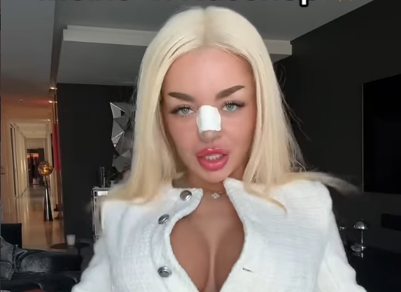 Blonde Frau mit Pflaster auf der Nase trägt eine weiße Jacke in einem modernen Raum.