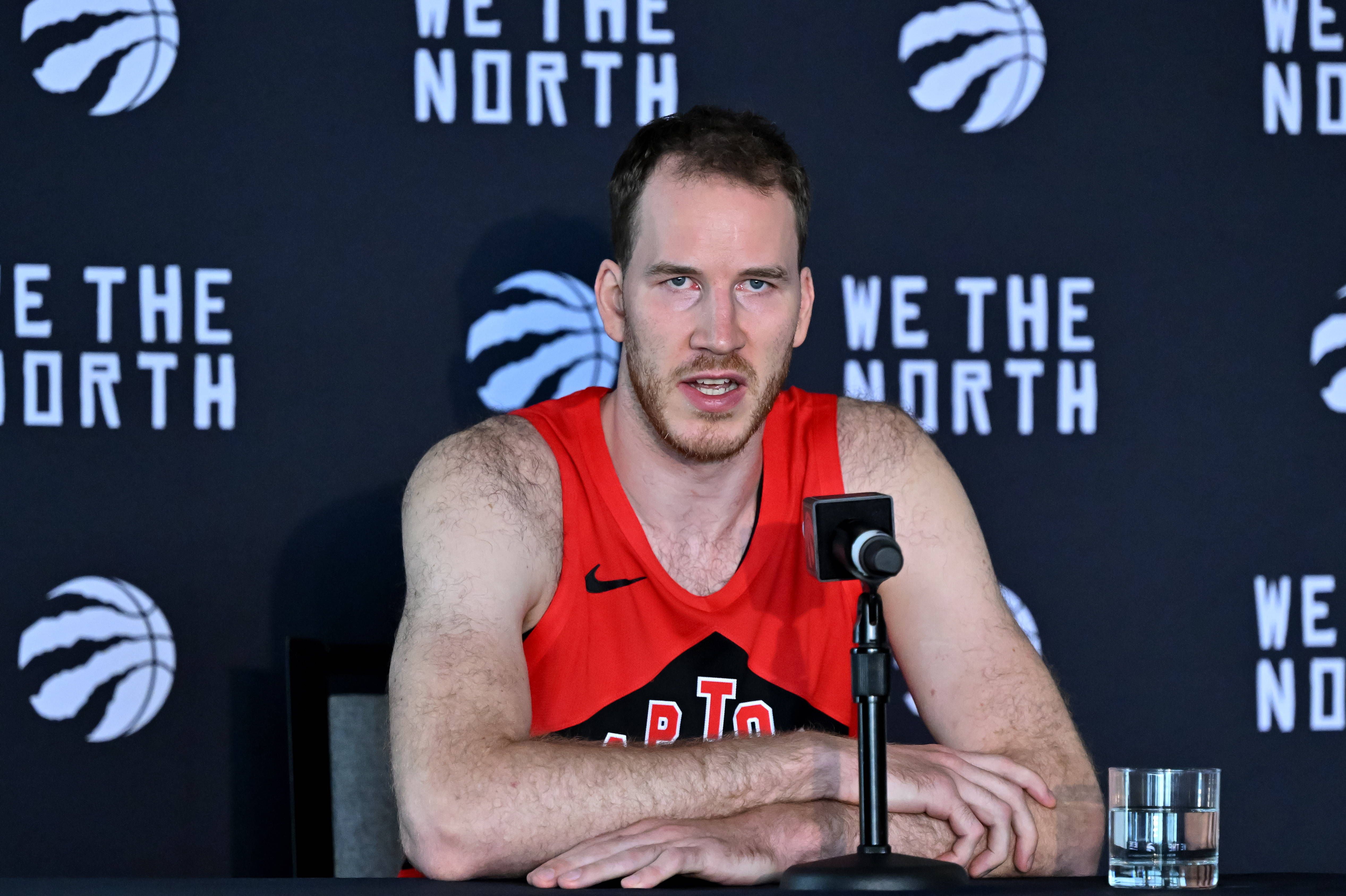 Jakob Poeltl spricht im Raptors-Trikot bei einer Pressekonferenz mit Mikrofon und Wasserglas.