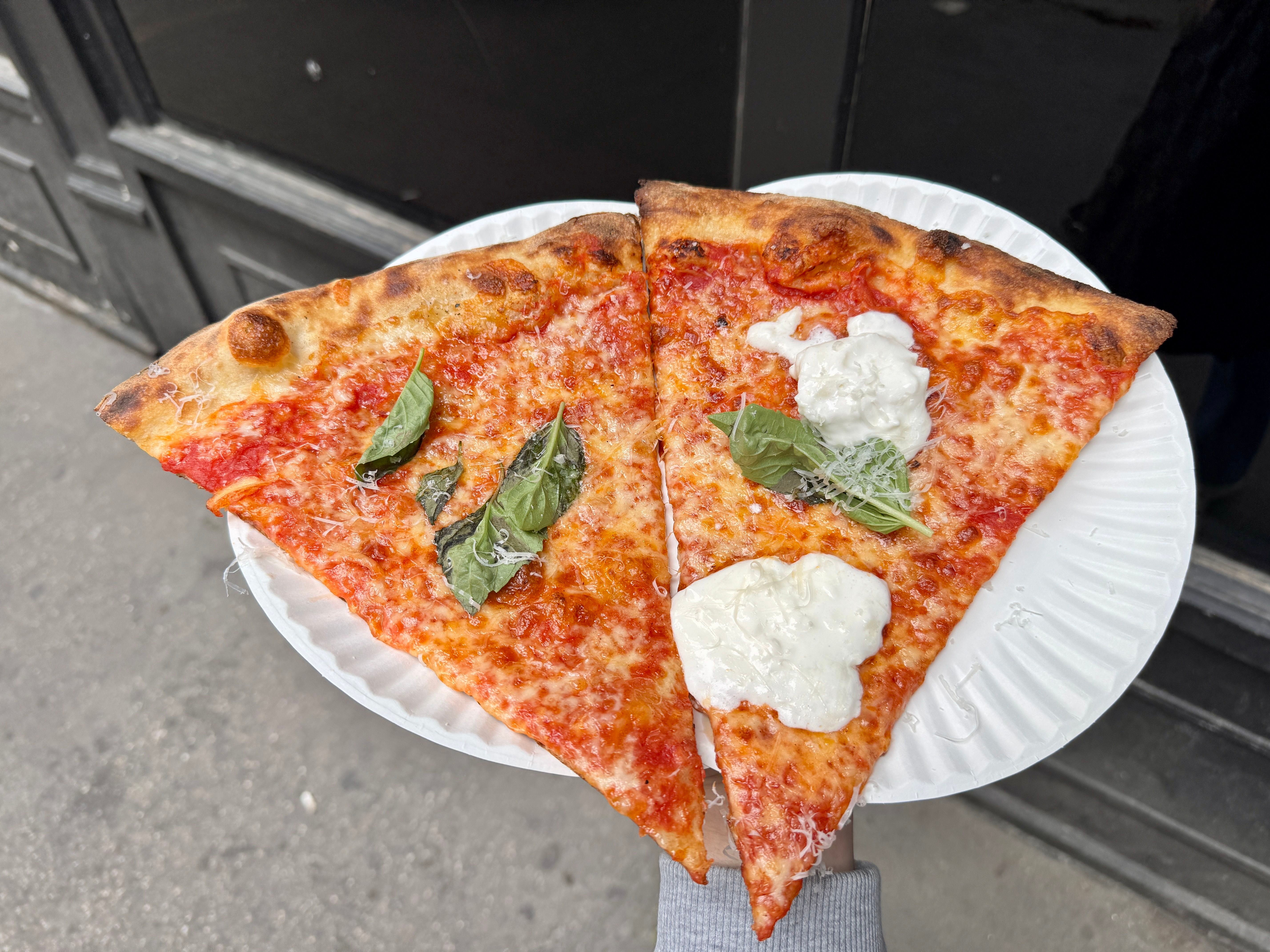 Zwei Pizzastücke mit Basilikum und Burrata auf einem Pappteller, auf einer Straße gehalten.