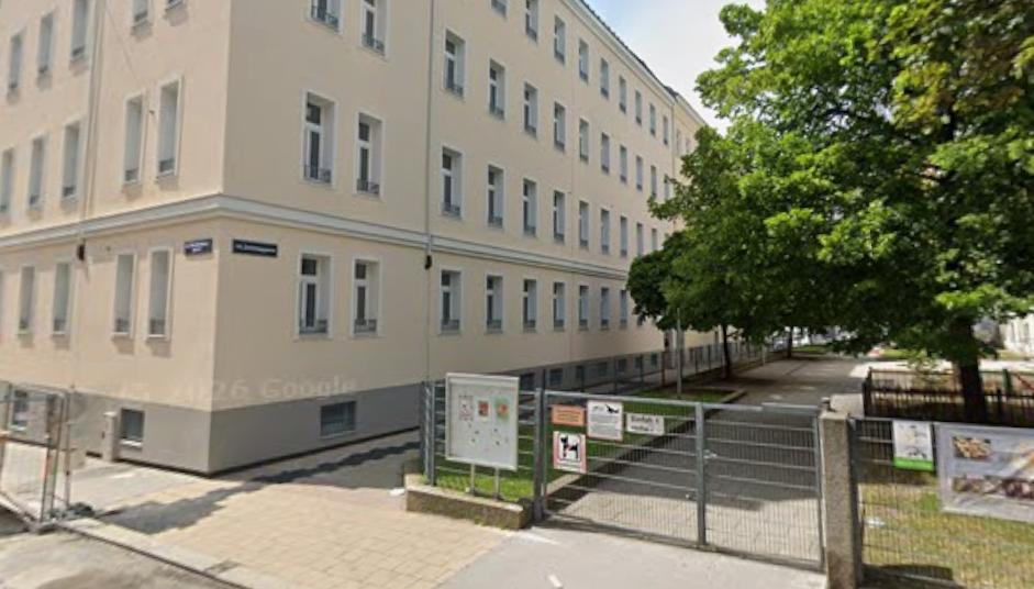 Gelbes Wohngebäude mit vielen Fenstern, Gittertor und Baum im Vordergrund in einer Stadtstraße.