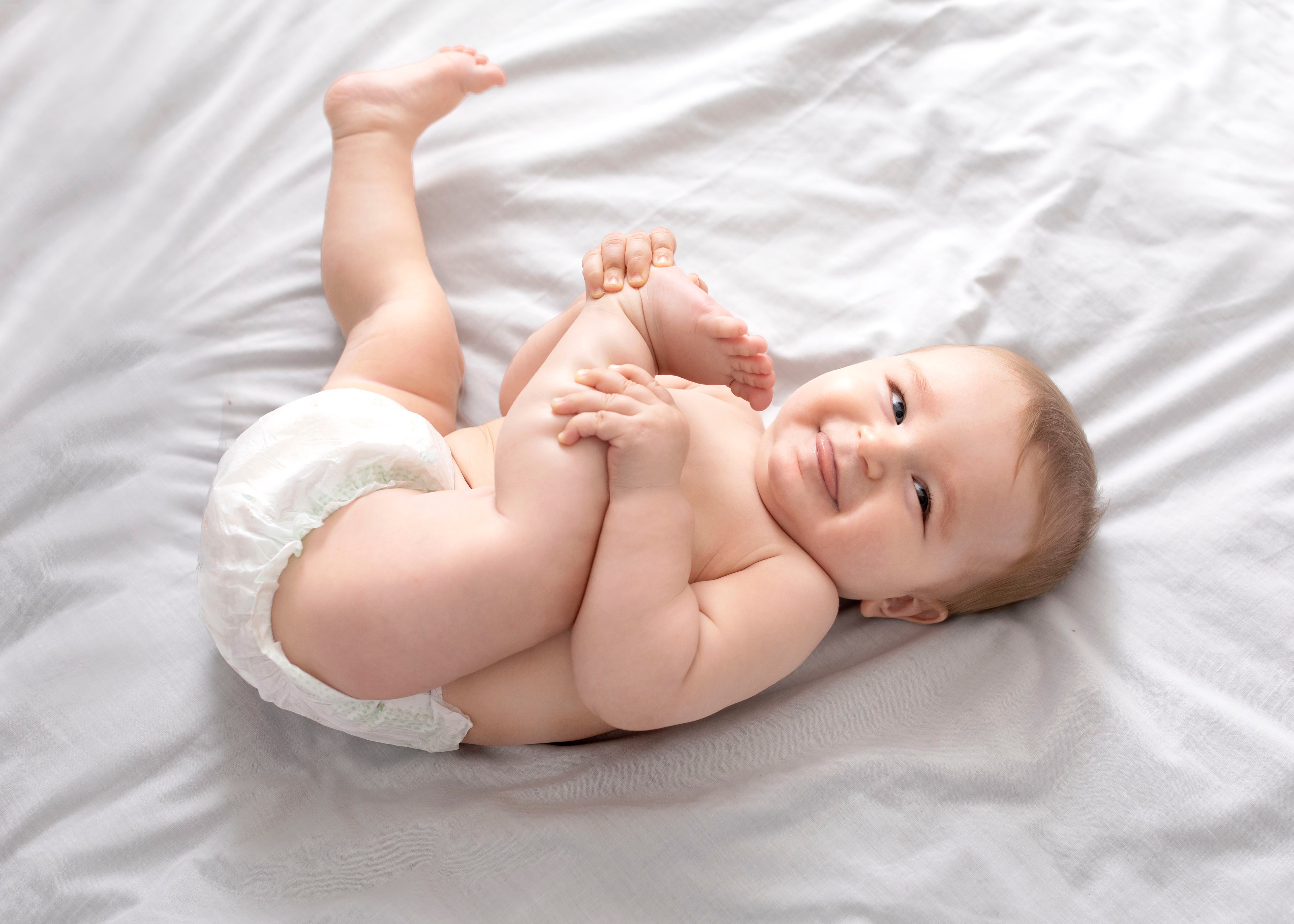 Lächelndes Baby in Windel liegt auf dem Rücken und spielt mit den Füßen auf weißem Bett.