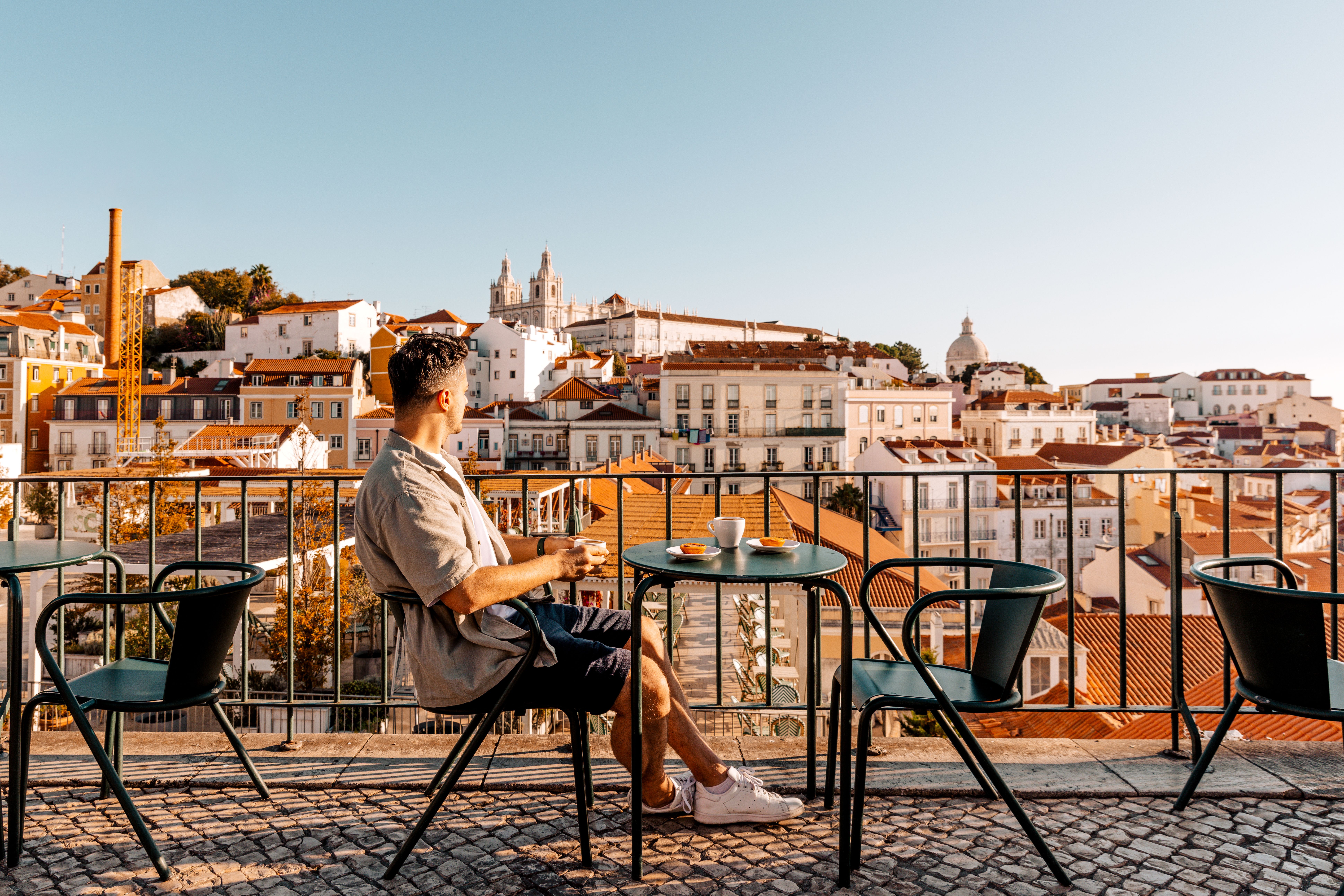Mann sitzt auf einer Terrasse mit Kaffee und Blick auf eine sonnige Stadt mit roten Dächern.