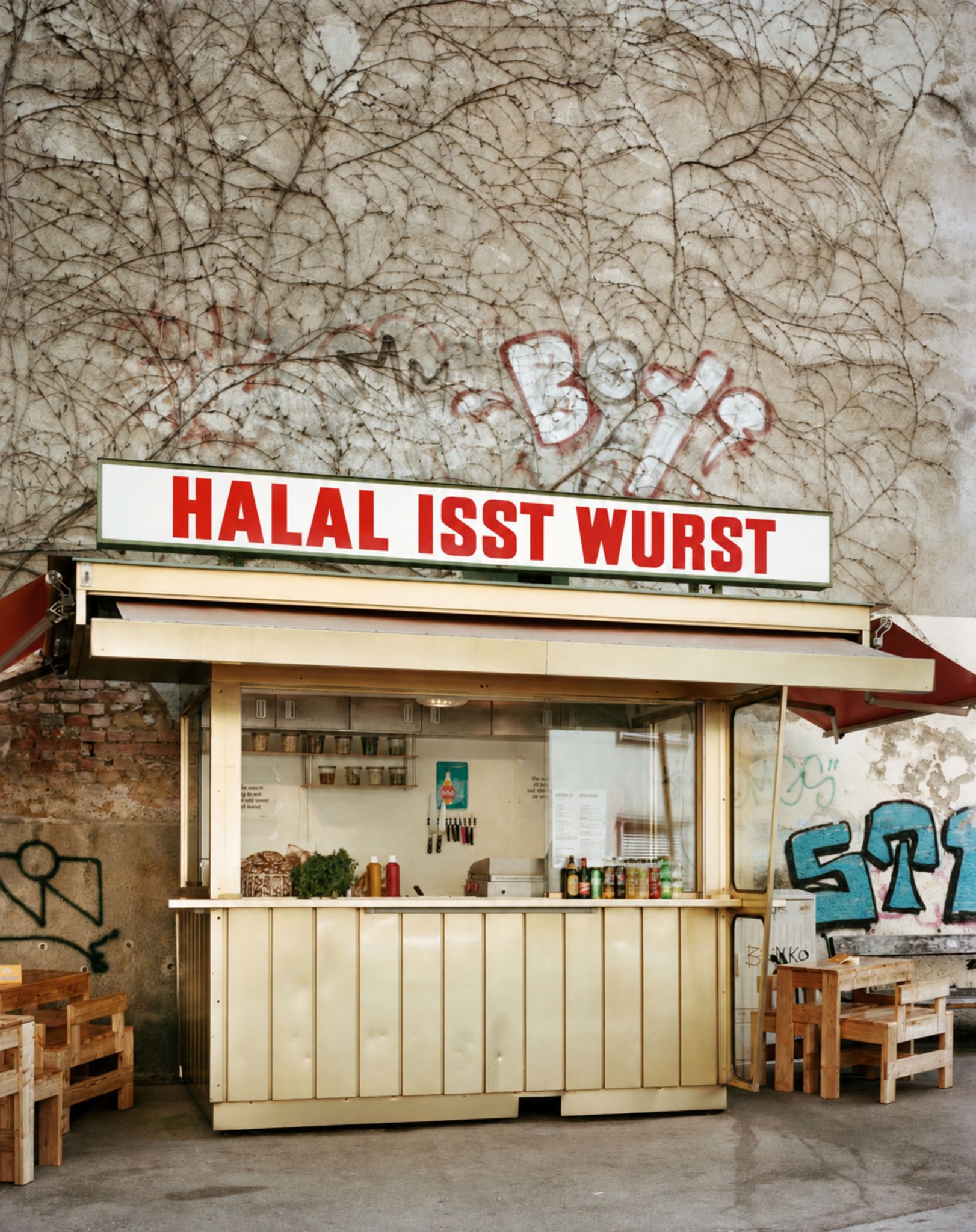 Kleiner Würstelstand mit Schild "HALAL ISST WURST" vor einer mit Graffiti besprühten Wand.