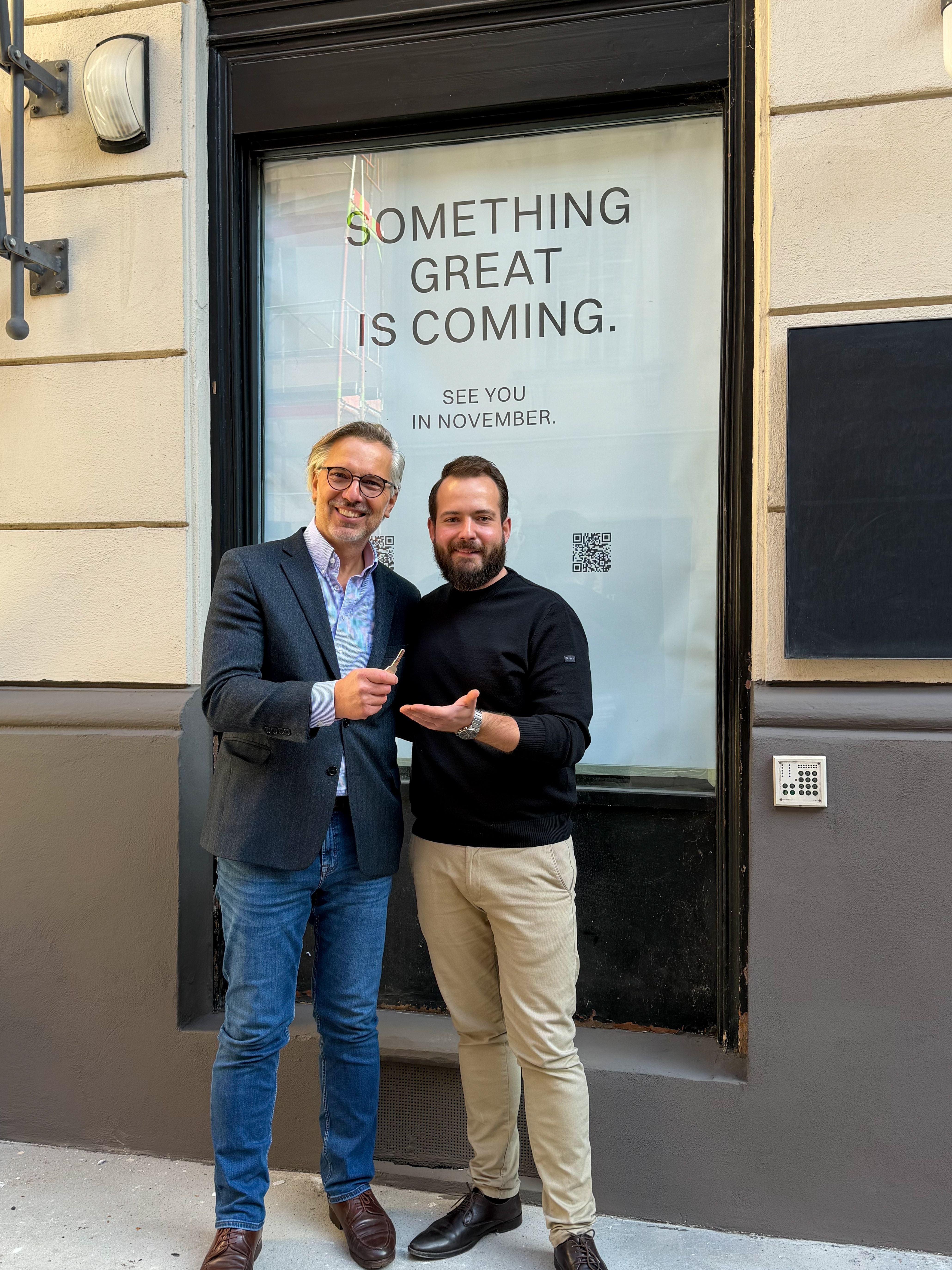 Zwei Männer stehen lächelnd vor einem Fenster mit dem Text „Something great is coming.“