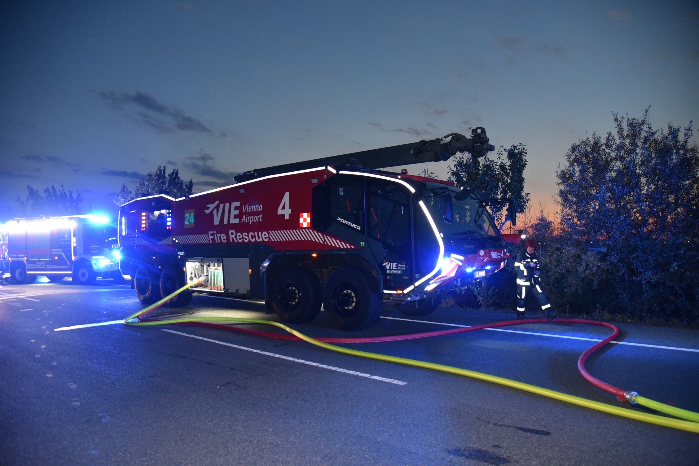 Feuerwehrfahrzeuge und Feuerwehrmann im Einsatz bei Dämmerung, Schläuche liegen auf der Straße.