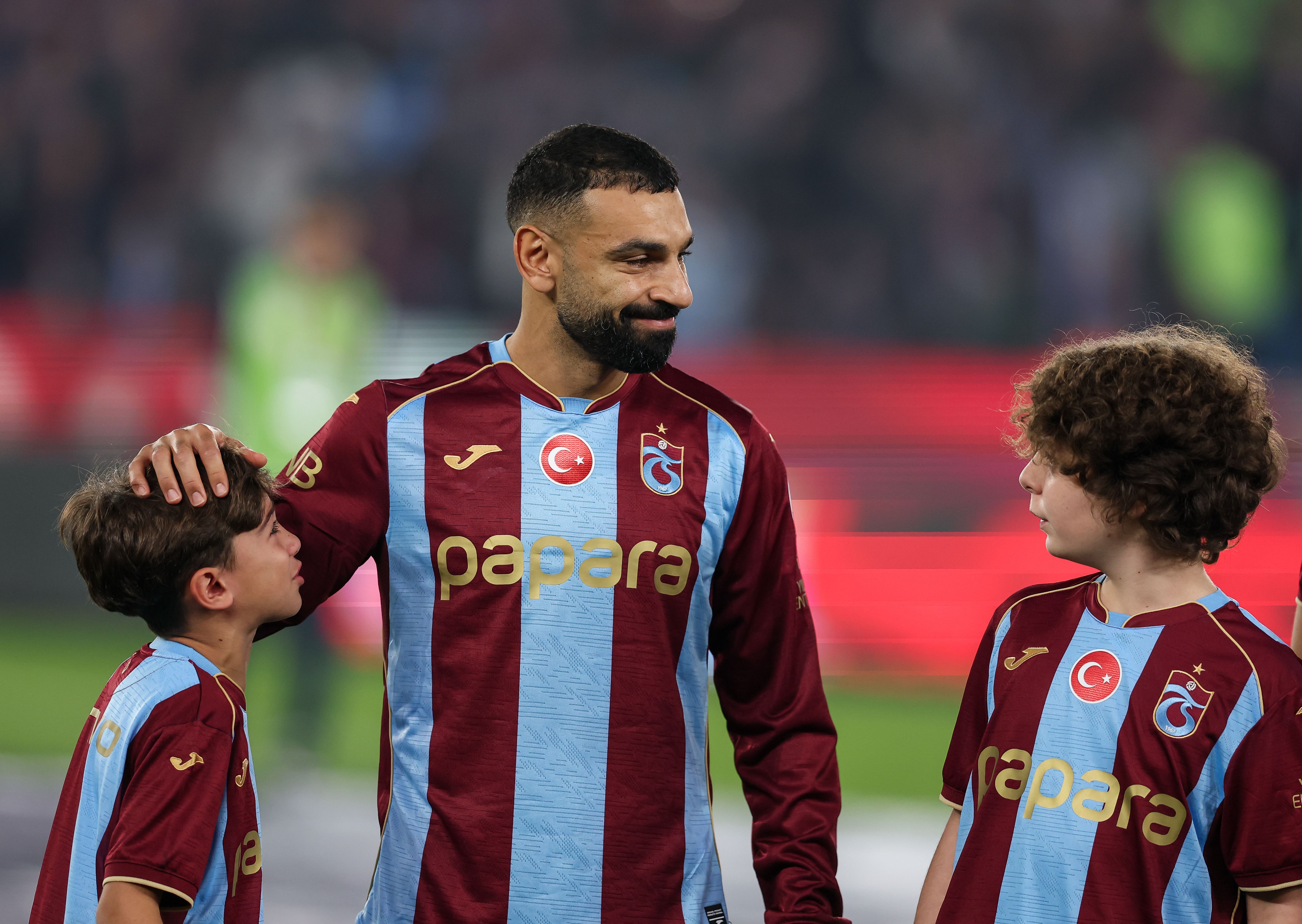 Mohamed Salah lächelt im rot-blau gestreiften Trikot von Trabzonspor auf dem Spielfeld und legt seine Hand sanft auf den Kopf eines jungen Einlaufkindes.