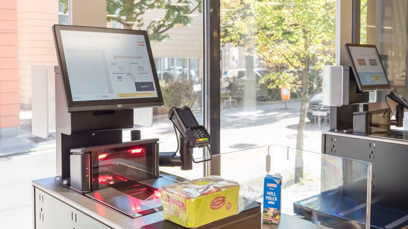 SB-Kasse bei Hofer mit Bildschirm, Scanner, Milch und einer Packung Toastbrot auf dem Kassenband.