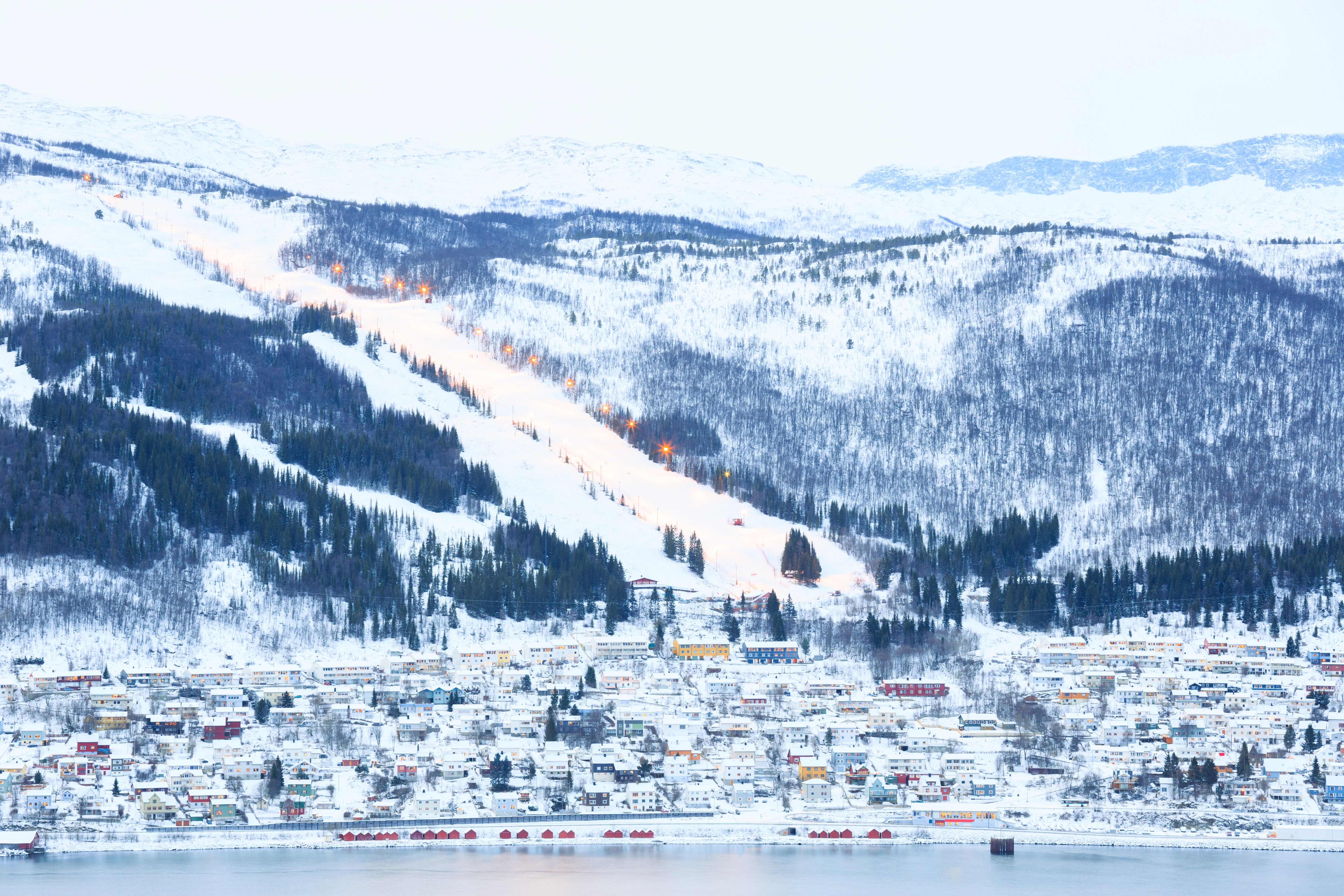 Verschneite Stadt Narvik mit beleuchteter Skipiste am Hang, umgeben von Bergen in Norwegen.