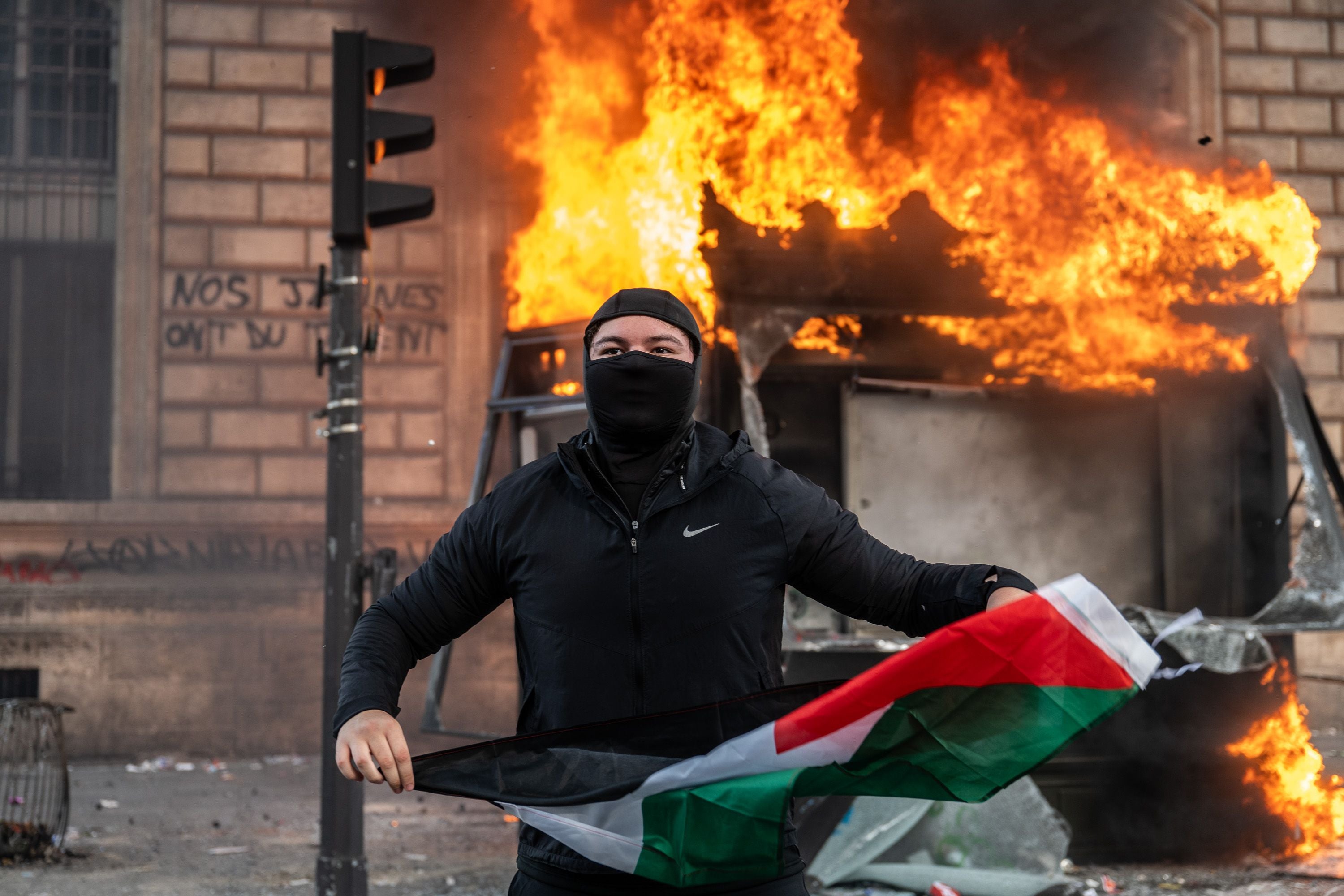 Ein vermummter Demonstrant hält eine Palästina-Flagge vor brennender Bushaltestelle in Paris.