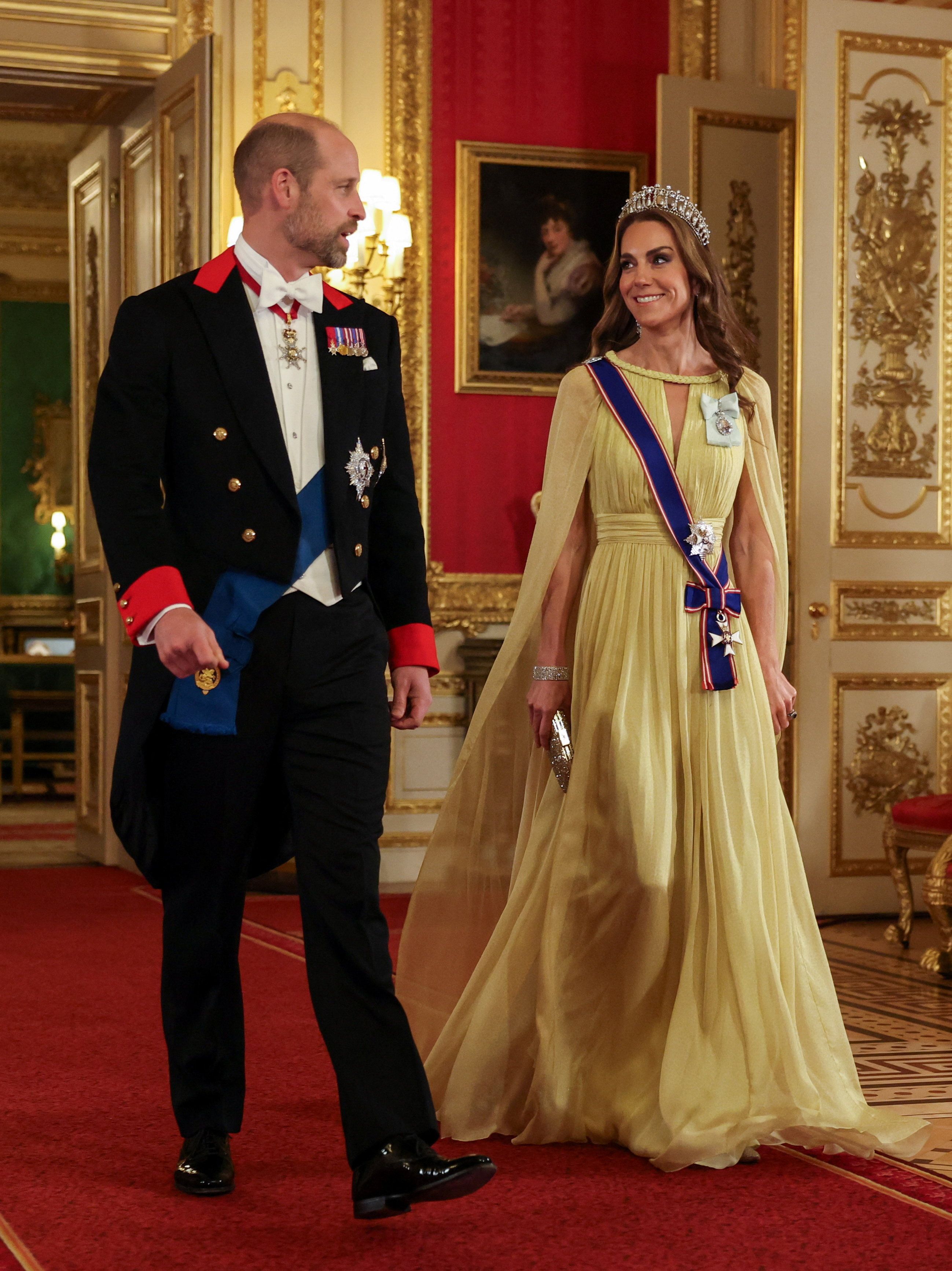 Ein Mann im Frack und eine Frau im gelben Abendkleid mit Tiara schreiten lächelnd durch einen prunkvollen Saal.