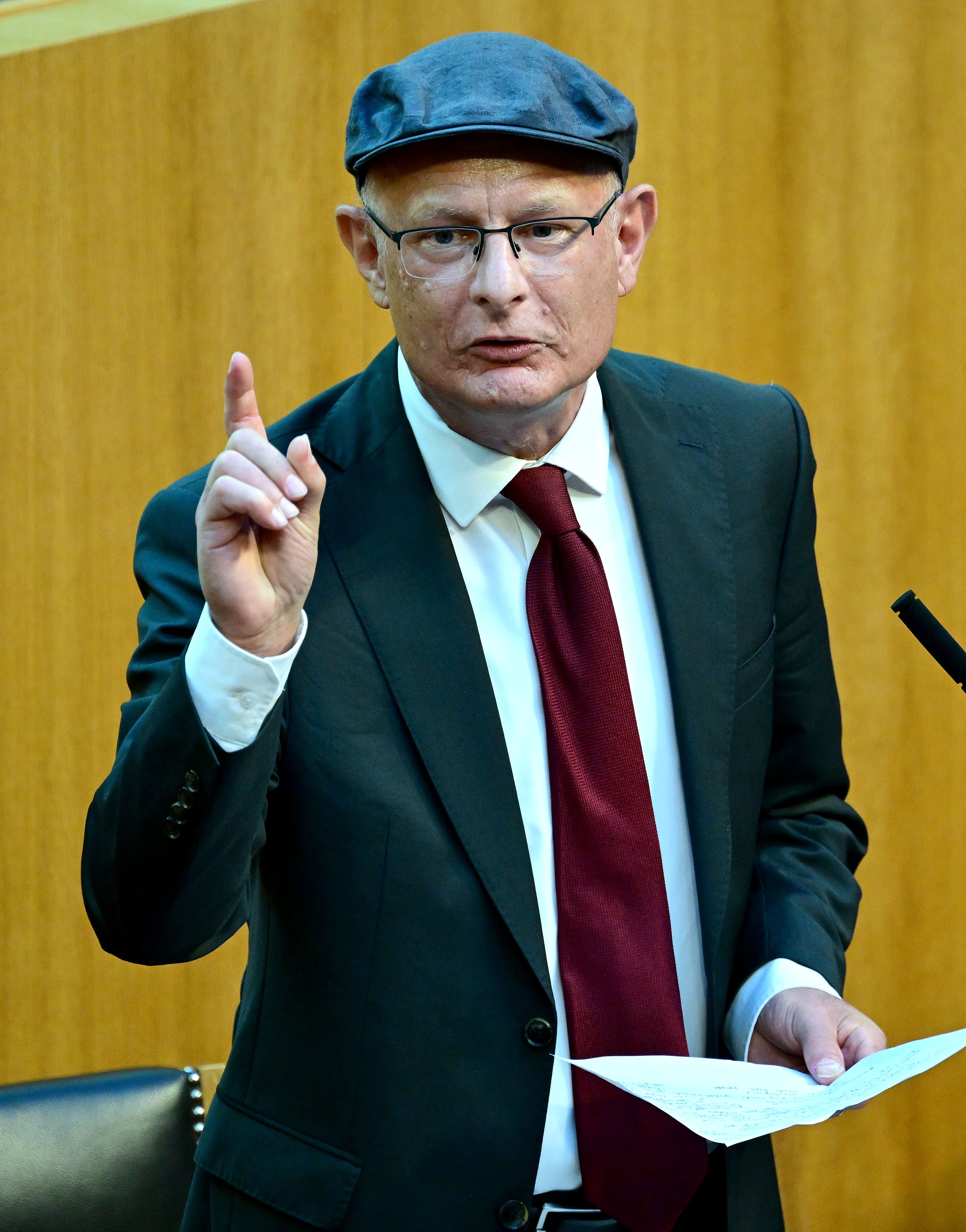 Ein Mann im Anzug mit roter Krawatte und Mütze spricht und gestikuliert im Parlament.
