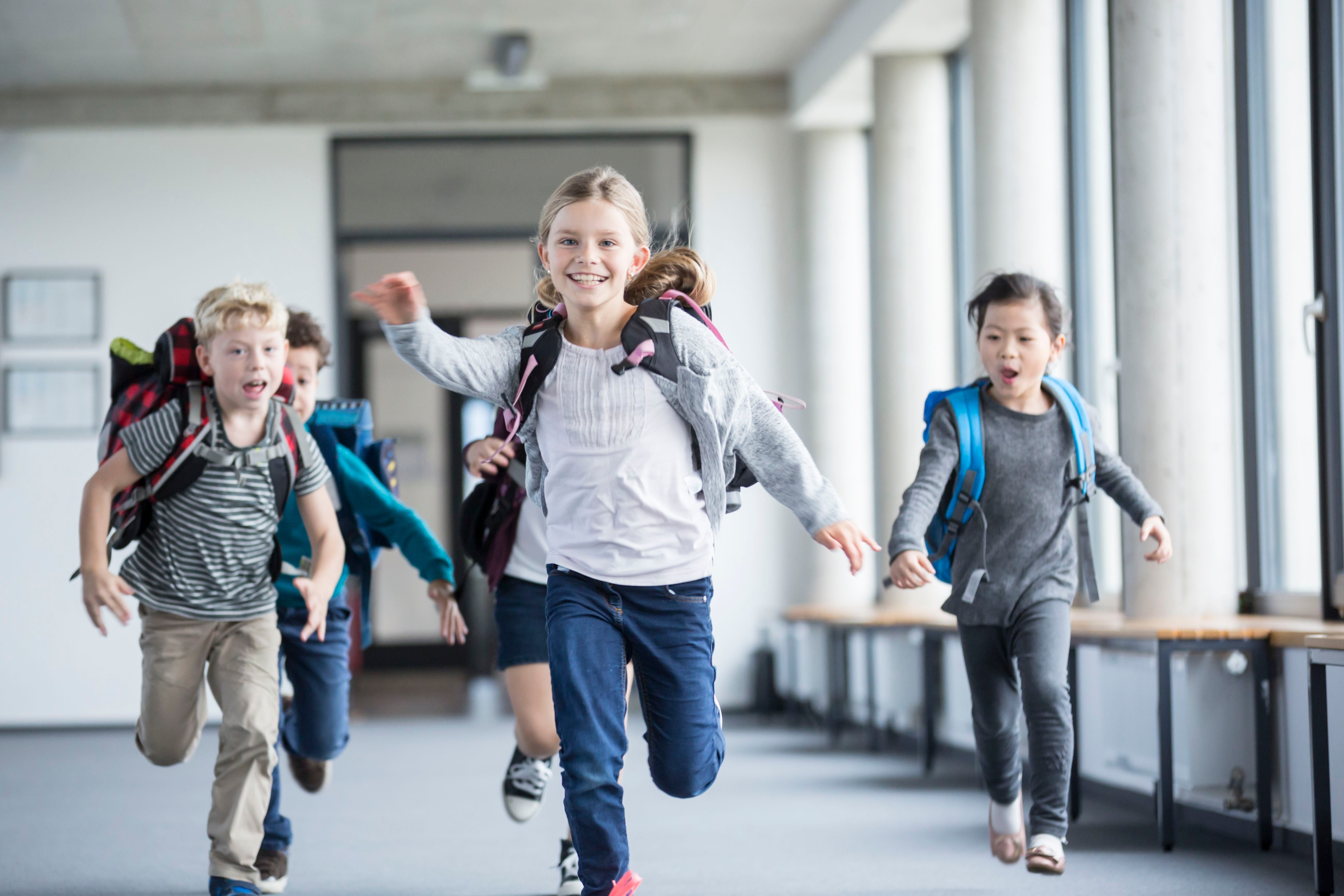 Vier Kinder mit Schulranzen rennen fröhlich durch einen hellen Schulflur.