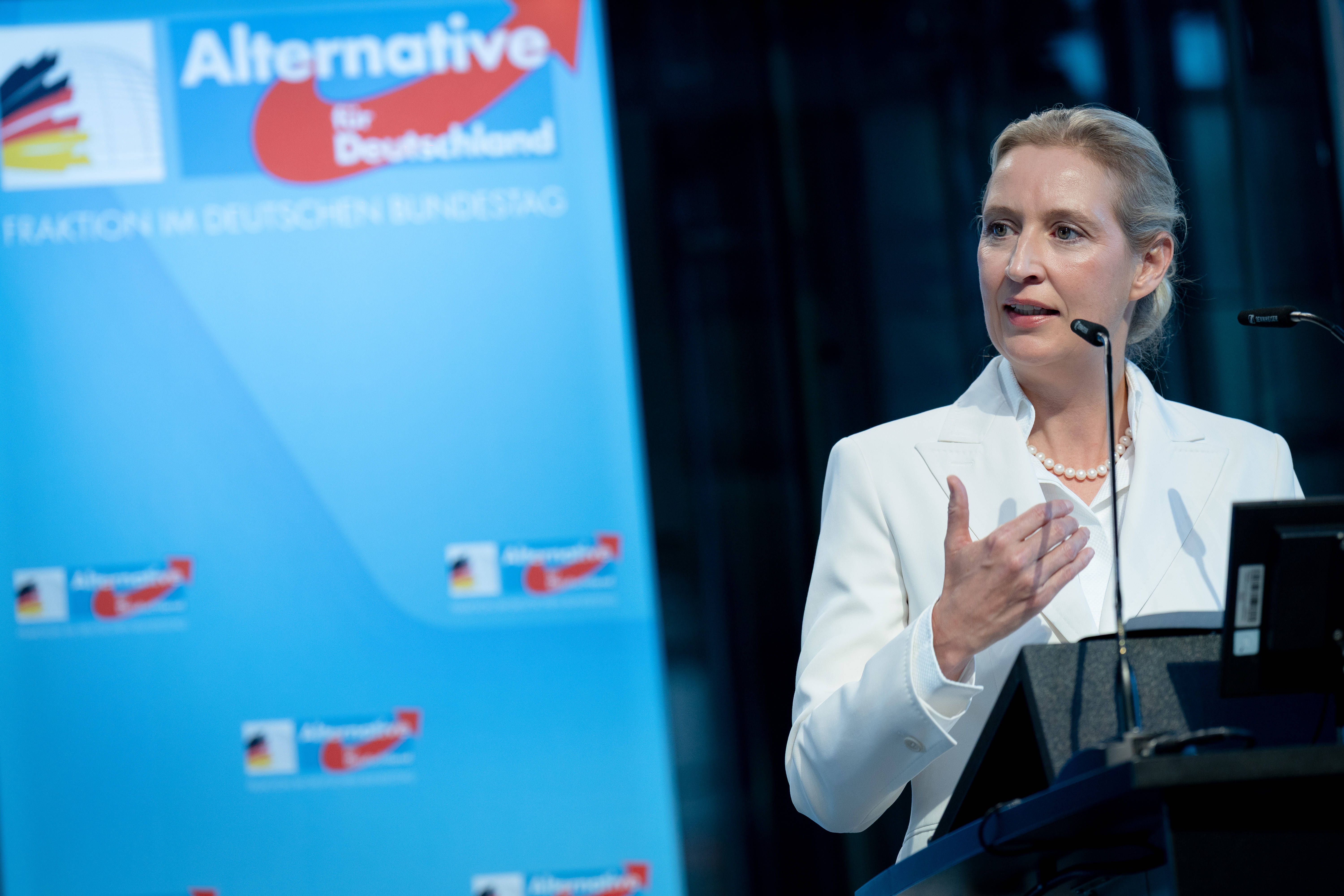 Eine Frau in weißem Anzug spricht am Rednerpult vor einem blauen AfD-Forum-Hintergrund.