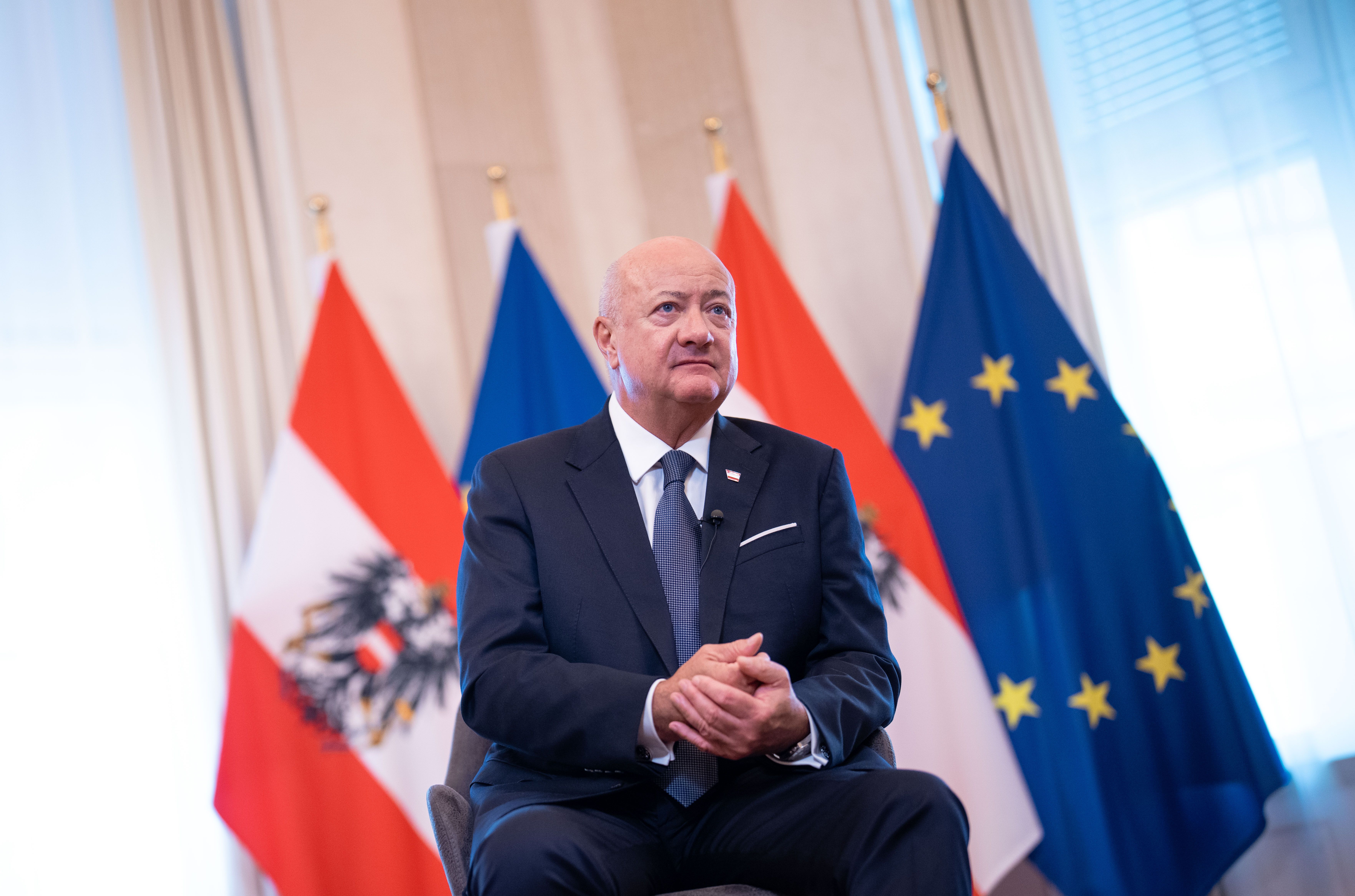 Ein Mann im Anzug sitzt vor österreichischen und EU-Flaggen in einem hellen Raum.