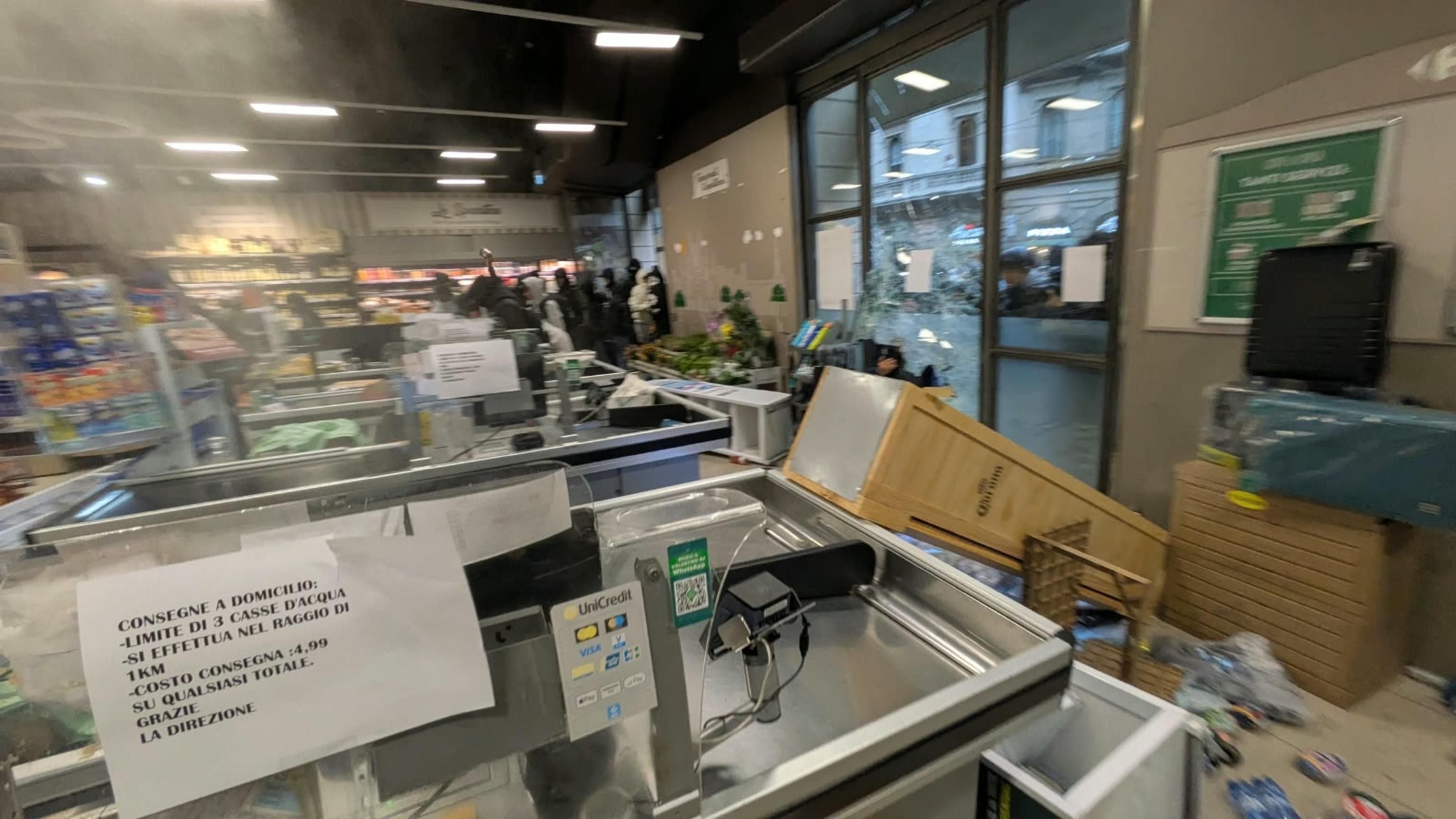 Verwüsteter Supermarkt nach Plünderung, gestürzte Regale und Unordnung, einige Menschen im Hintergrund.