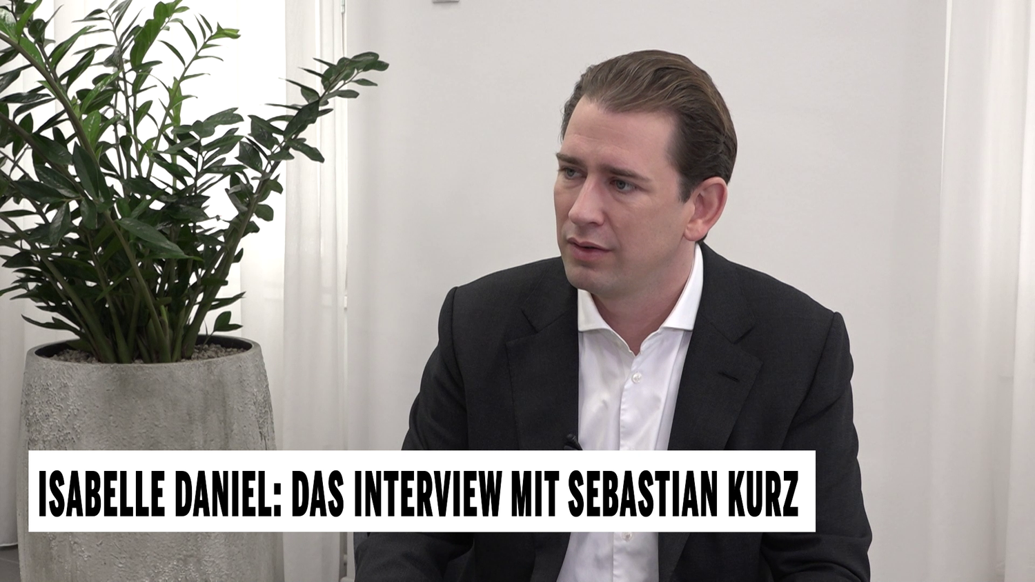 Ein Mann im Anzug sitzt bei einem Interview, im Hintergrund eine große Zimmerpflanze.