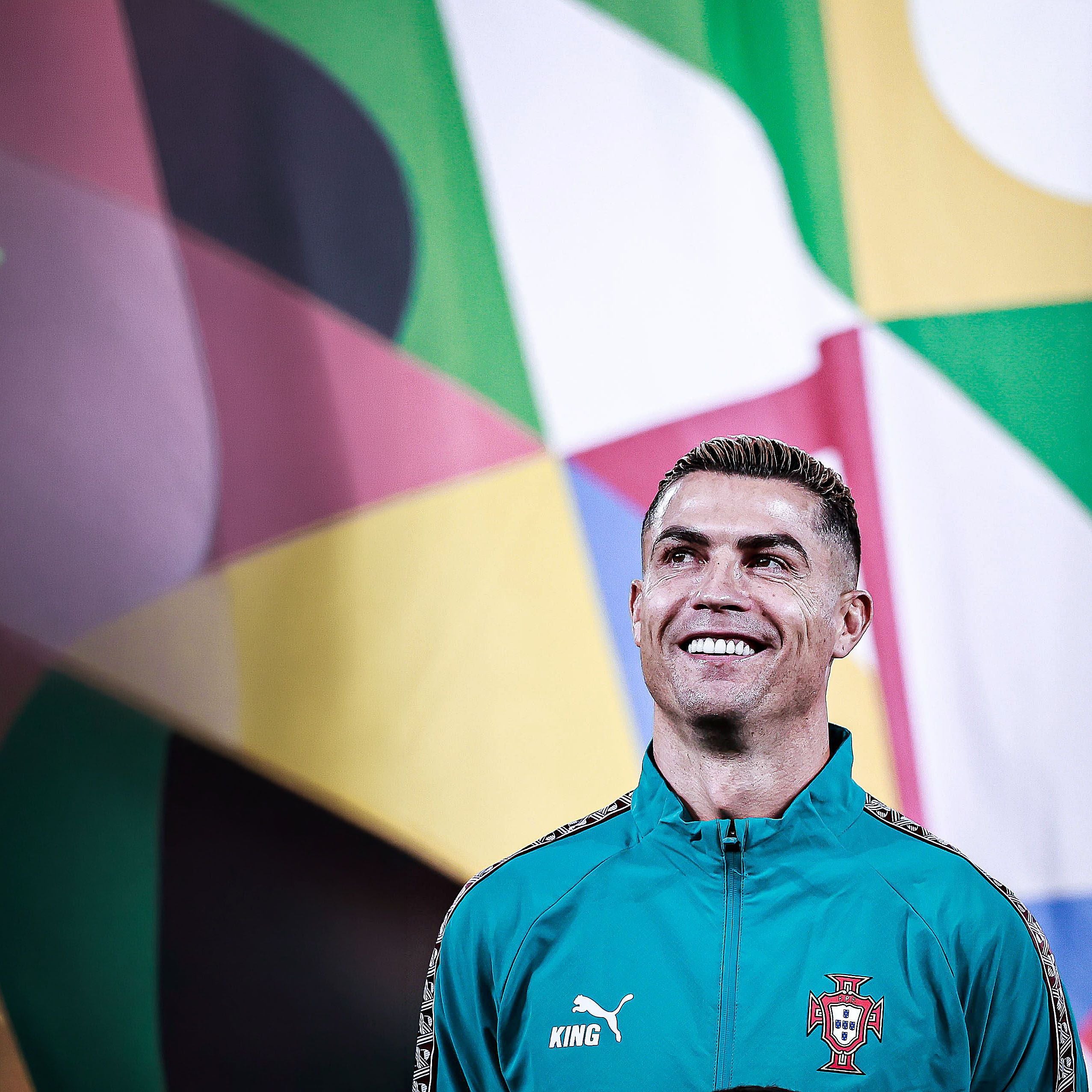 Cristiano Ronaldo lächelt zufrieden im türkisgrünen Trainingsanzug und blickt leicht nach oben vor buntem Hintergrund.