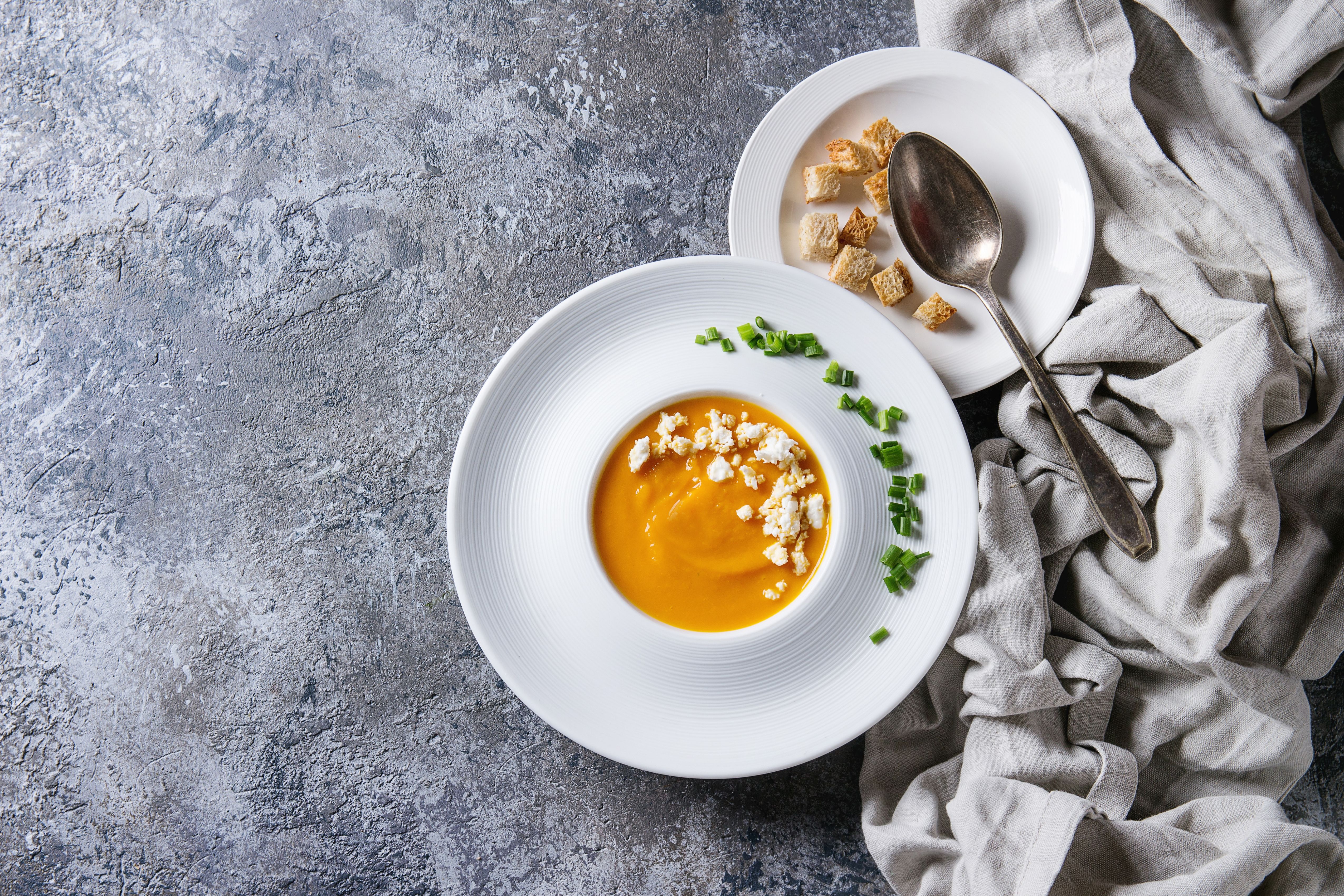 Teller mit Kürbis-Karottensuppe, Feta, Schnittlauch und Croutons, daneben Löffel auf Serviette.