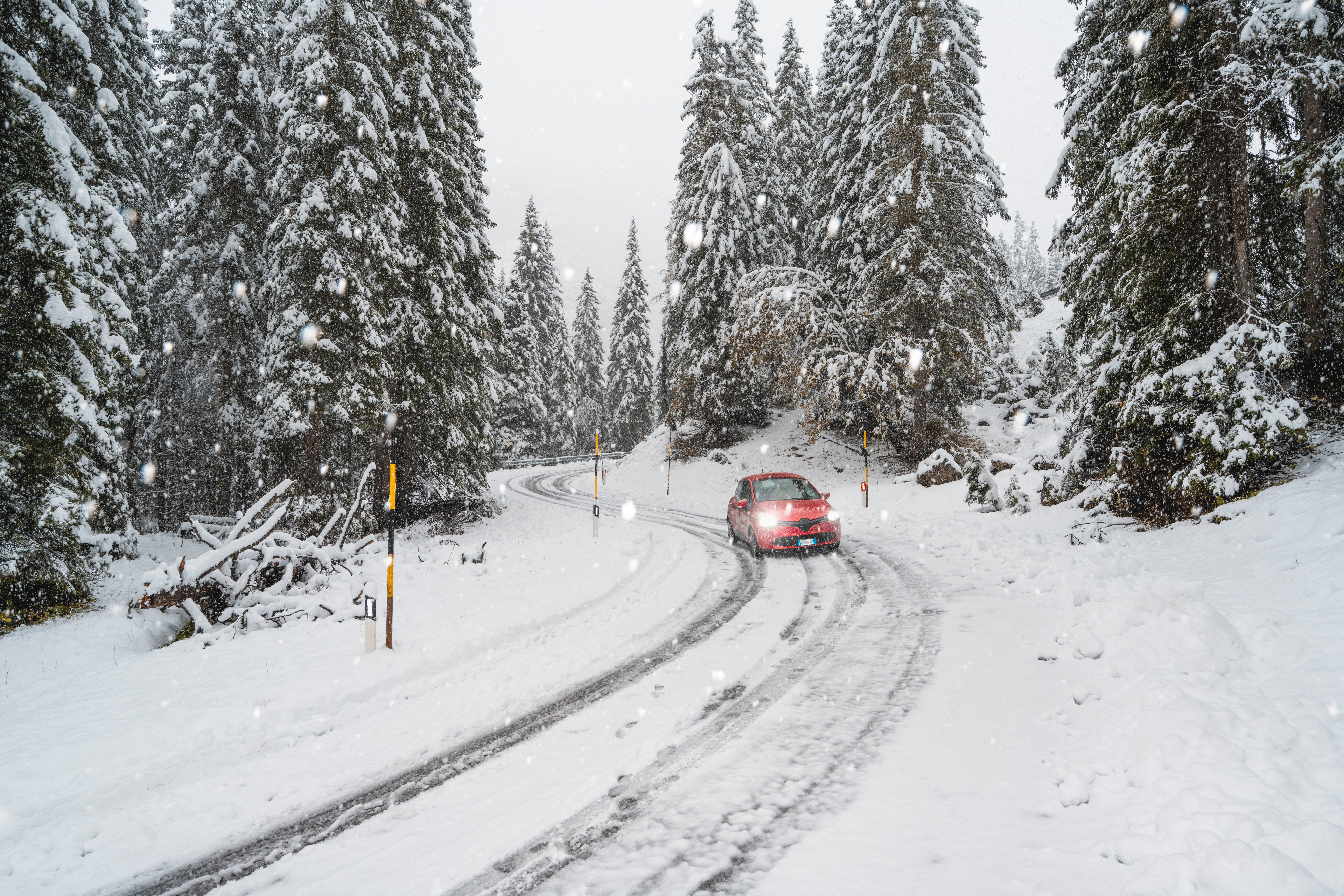 Rotes Auto fährt auf verschneiter Straße durch winterlichen Wald bei starkem Schneefall.