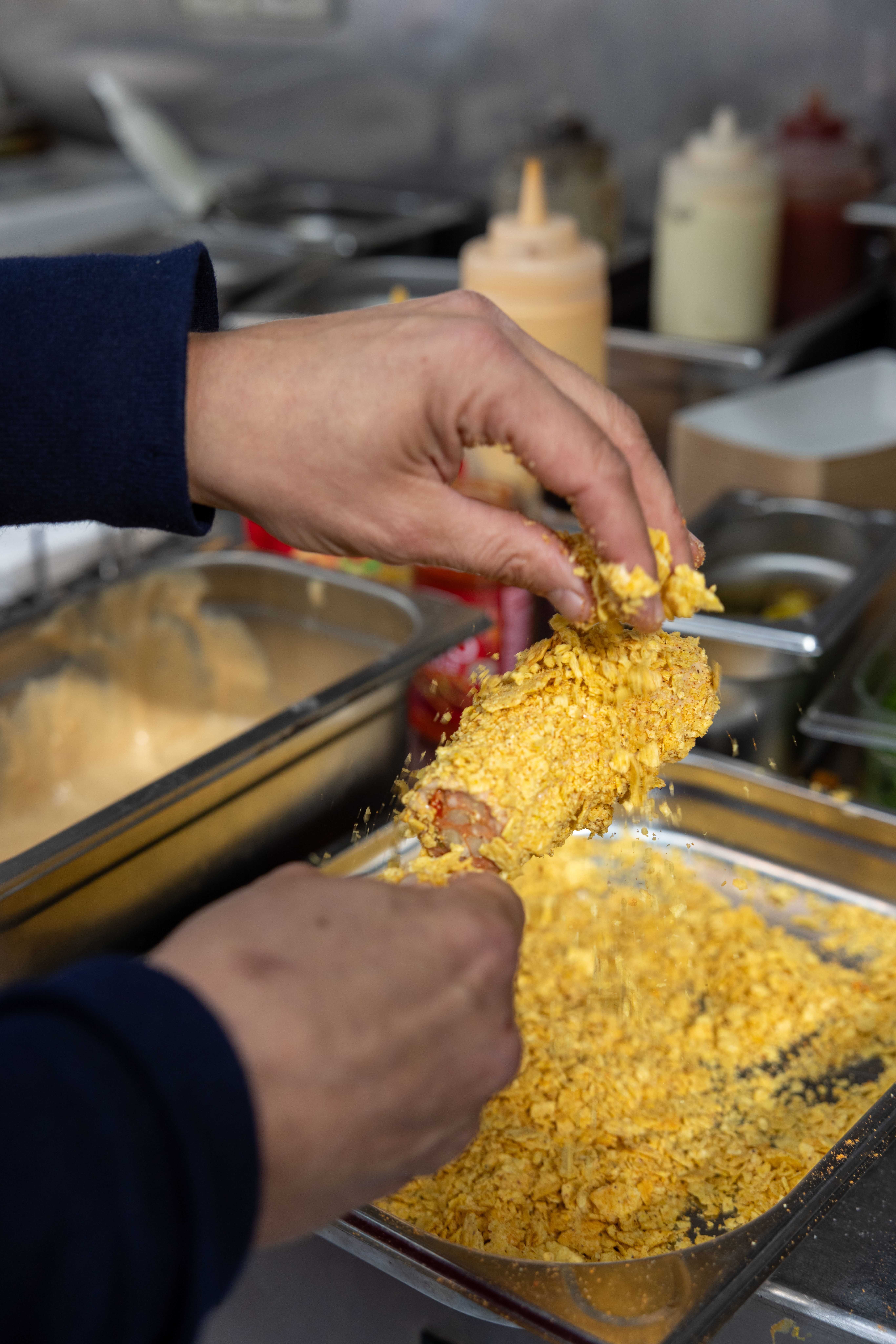 Eine Hand paniert einen Hotdog mit zerbröselten Nachochips in einer Gastronomie-Küche.