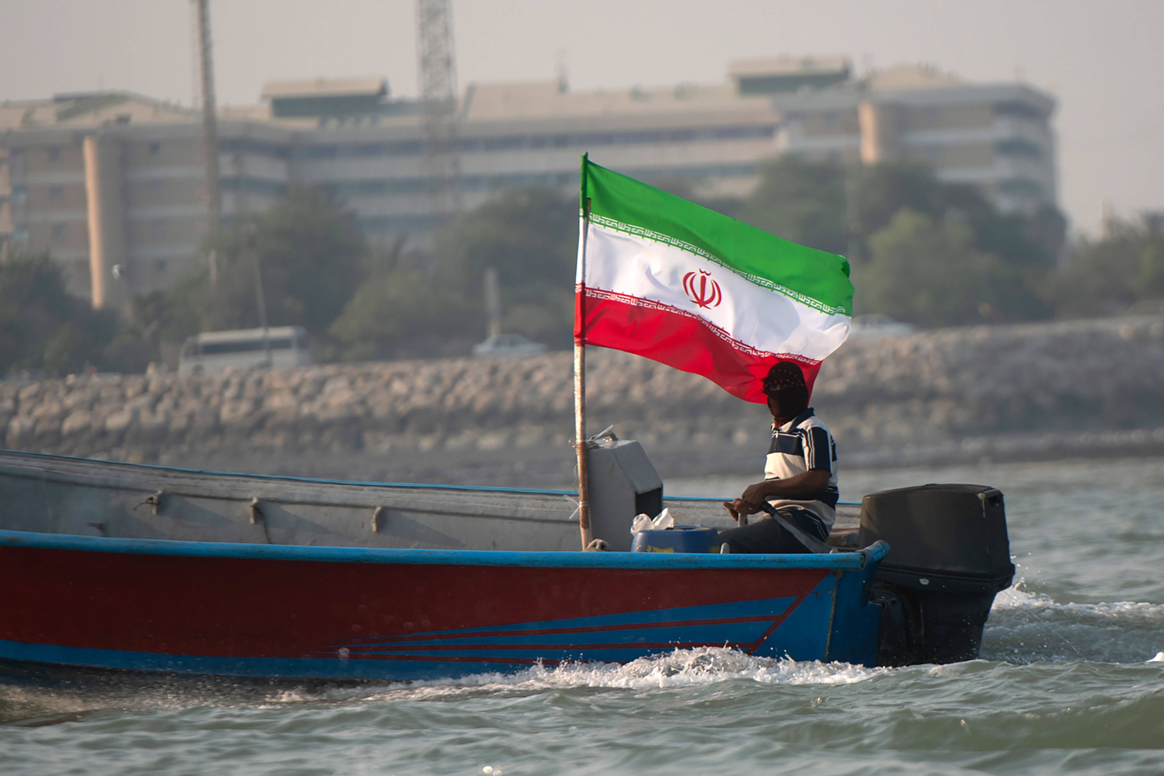 Ein Mann fährt ein Boot mit iranischer Flagge entlang der Küste, Gebäude im Hintergrund.