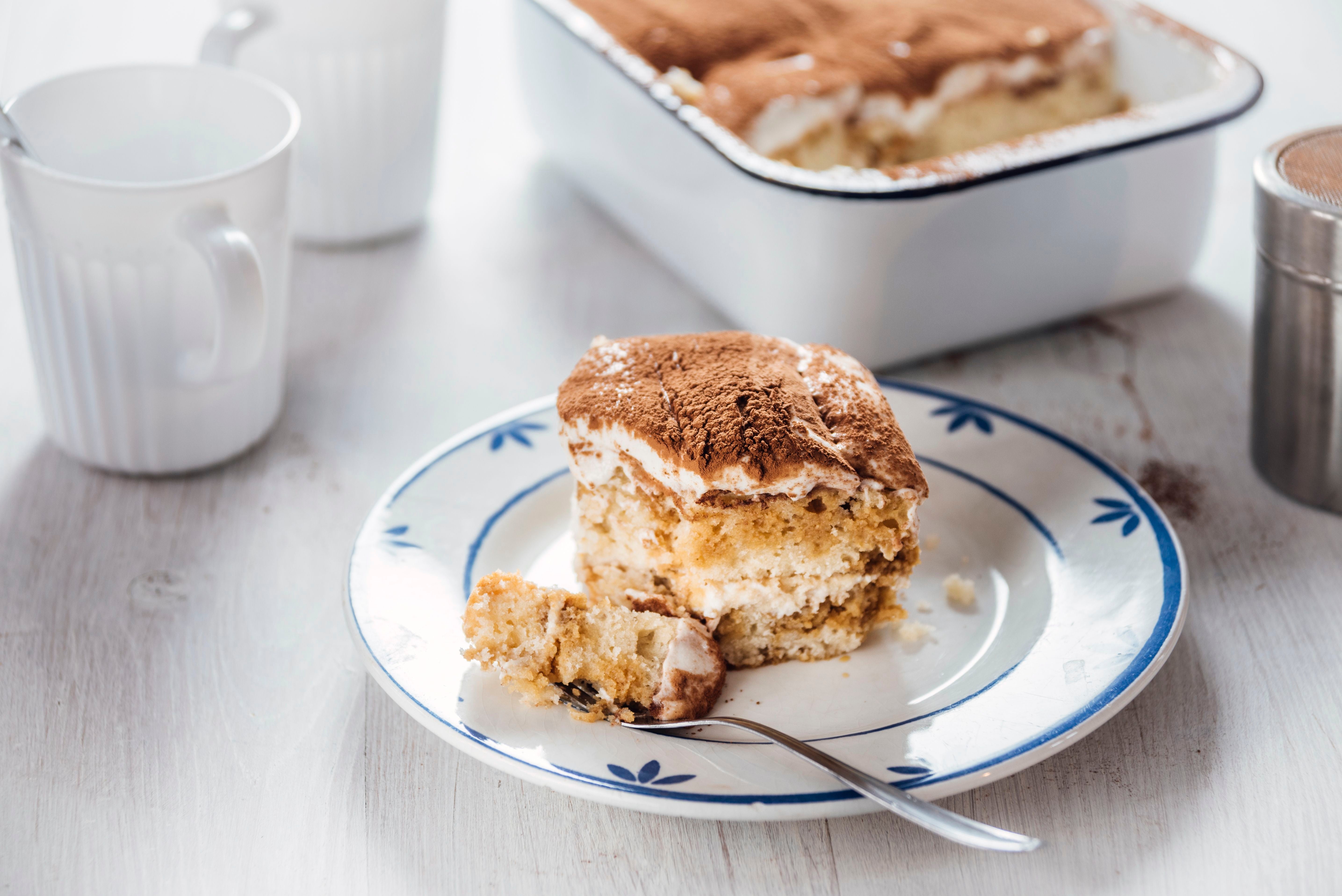 Ein Stück Tiramisu liegt auf einem Teller, daneben ein Löffel und Tassen im Hintergrund.