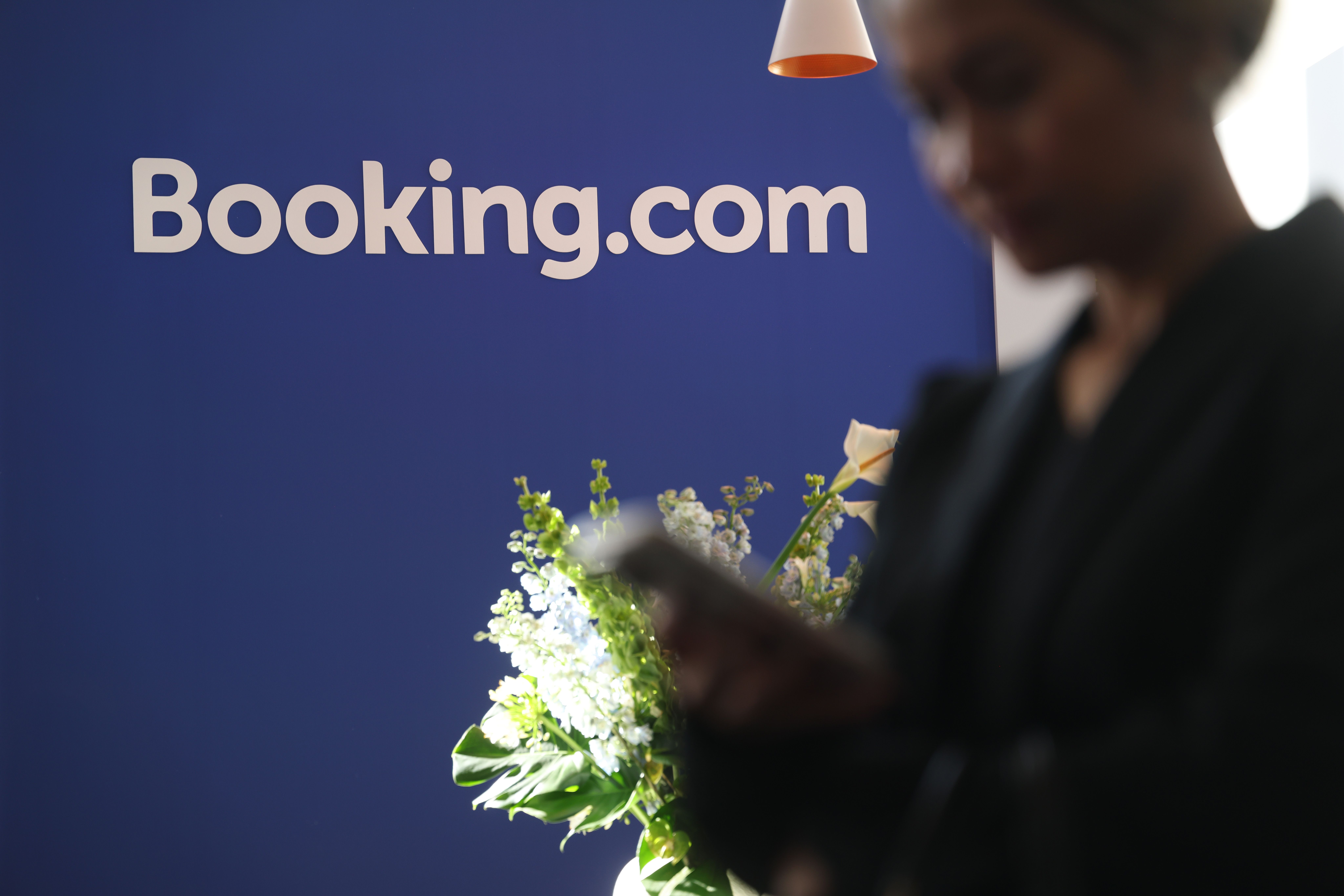 Eine Person benutzt ein Smartphone vor einem Booking.com-Stand mit Blumenarrangement.